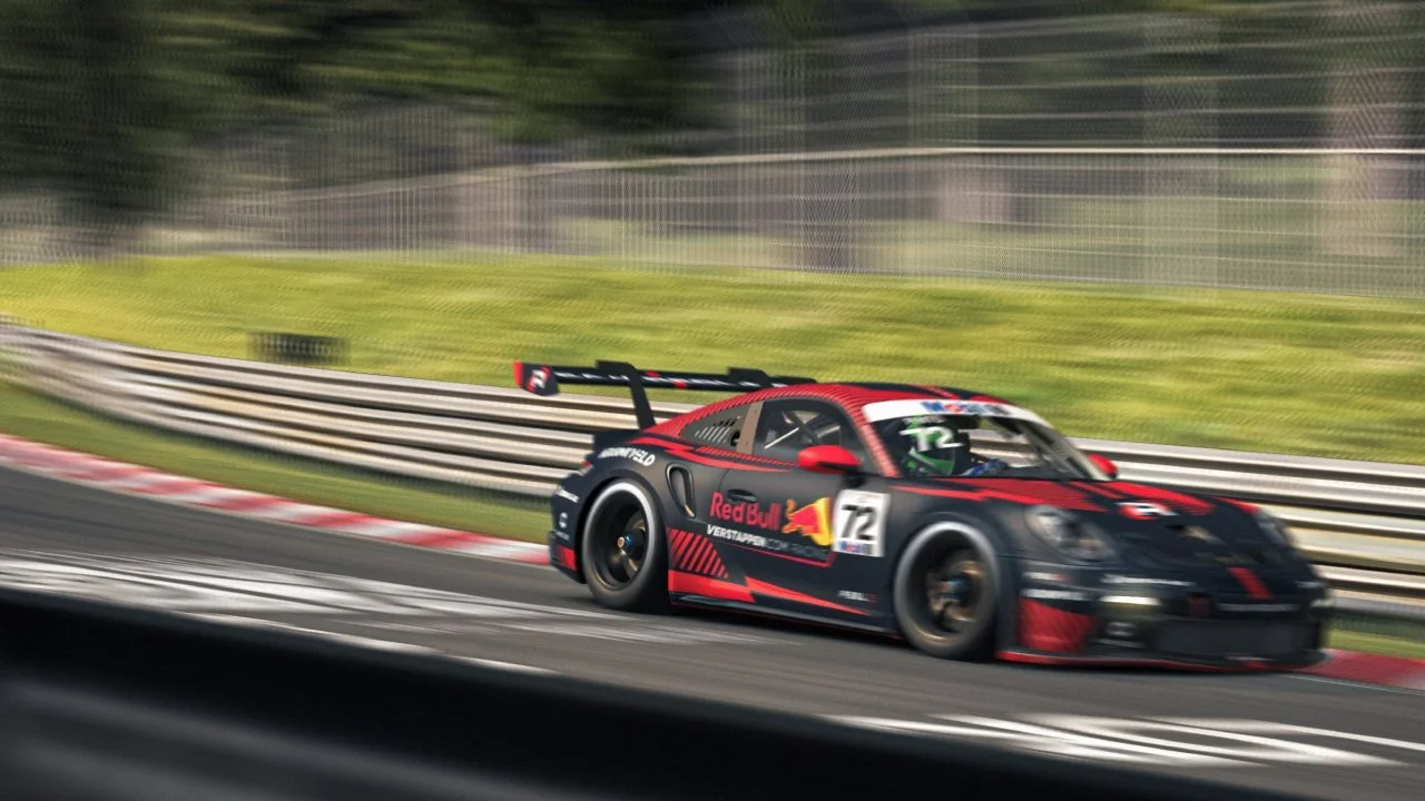 A red and black sports car with a wing spoiler races on a track, featuring Red Bull and Verstappen branding. The background shows blurred greenery and a fence, indicating high speed.