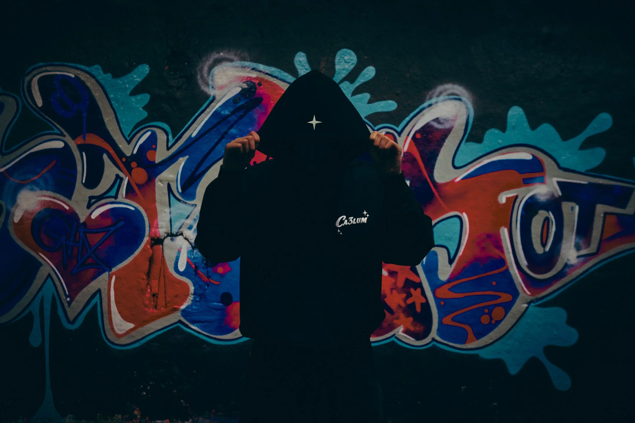 Person wearing a hooded jacket with a star on the hood, standing in front of a colorful graffiti wall, with their face obscured by the hood.