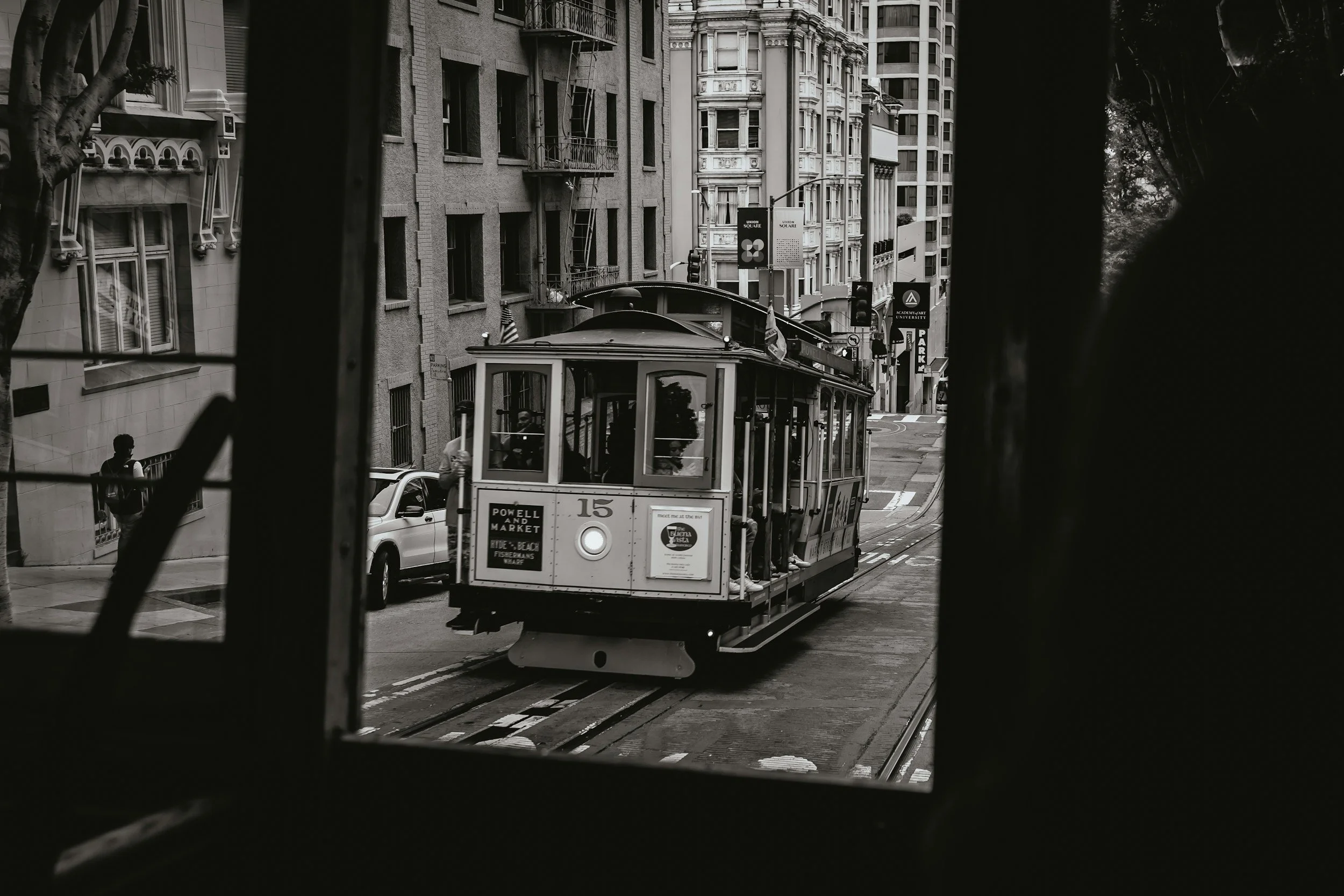 A vintage San Francisco cable car traveling up a city street with buildings on either side, viewed through a window frame in the foreground.