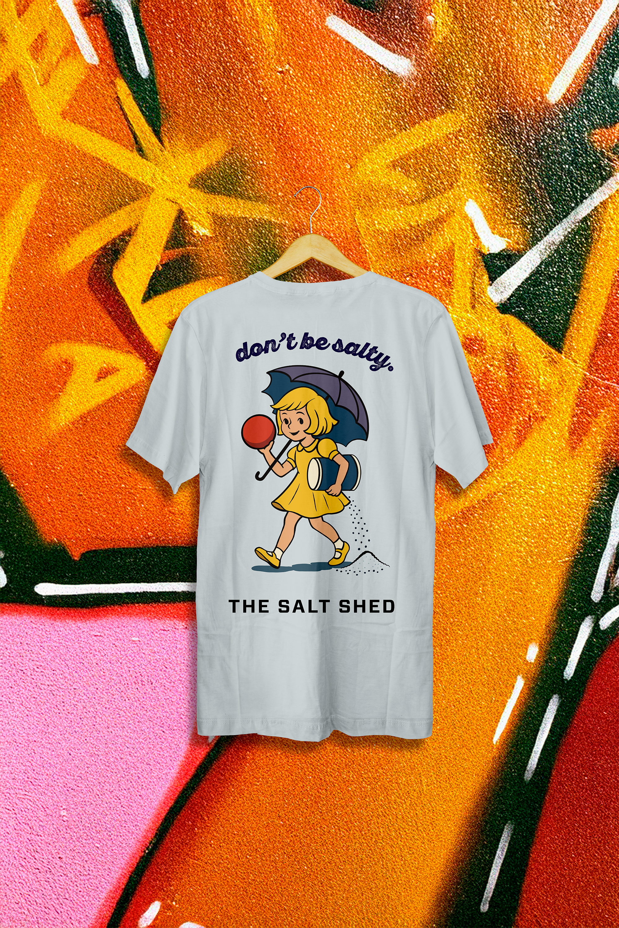 A white T-shirt hanging on a wooden hanger with a cartoon girl holding an umbrella and a bowling ball, and the text "don't be salty. THE SALT SHED". The background features brightly colored graffiti wall art with orange, pink, green, and black accents.