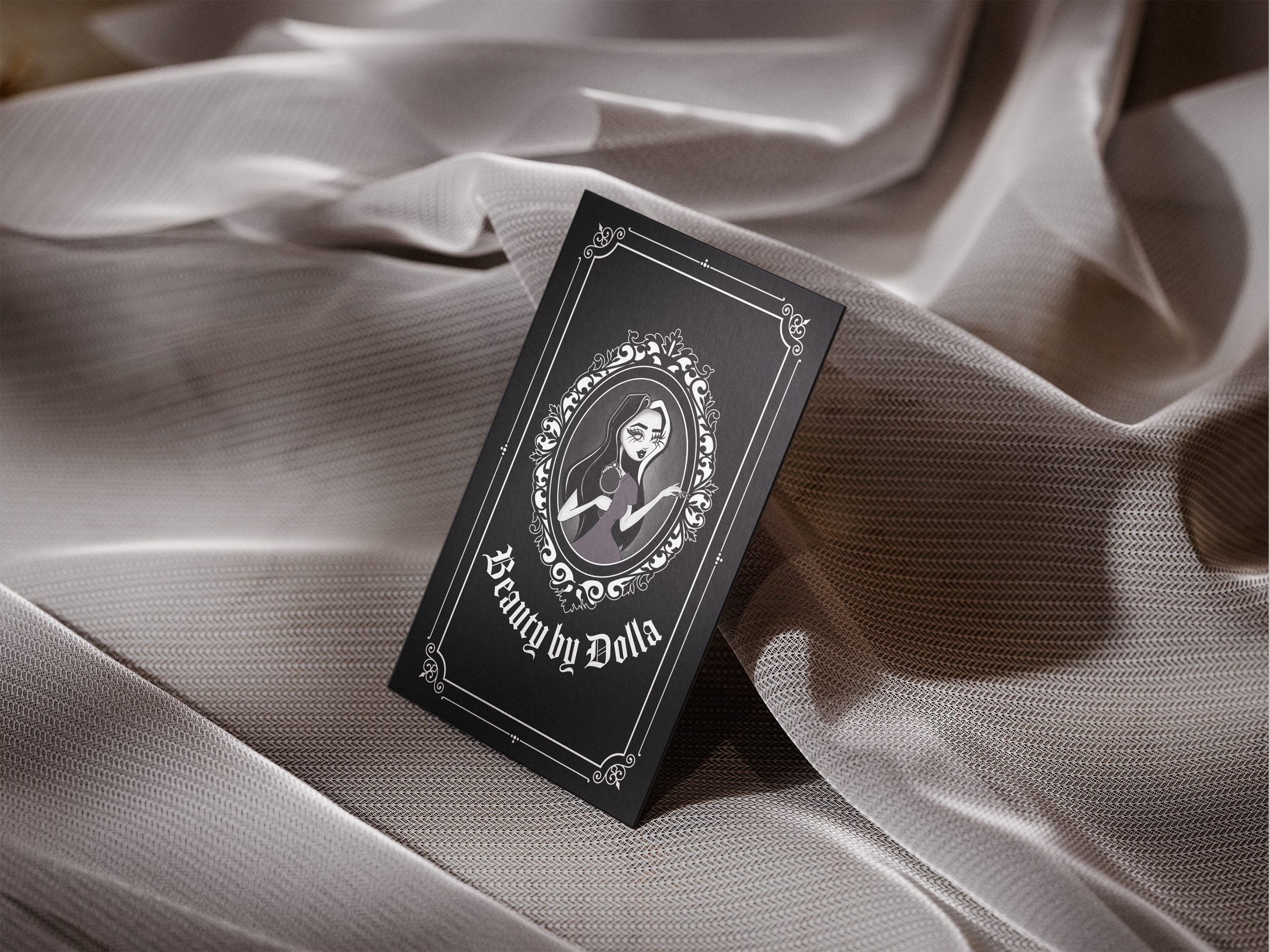 A black business card with a white illustration of a woman holding a cigarette, surrounded by an ornate oval frame, with the text "Beauty by Dolla" underneath, placed on soft, beige satin fabric.