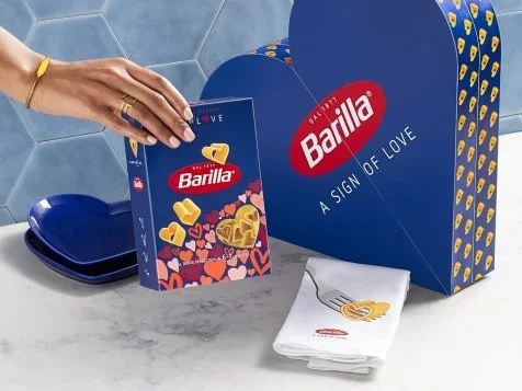 A hand holding a Barilla pasta box next to a blue heart-shaped gift box with Barilla branding, a white cloth with a gold heart design, and blue plates on a kitchen countertop.