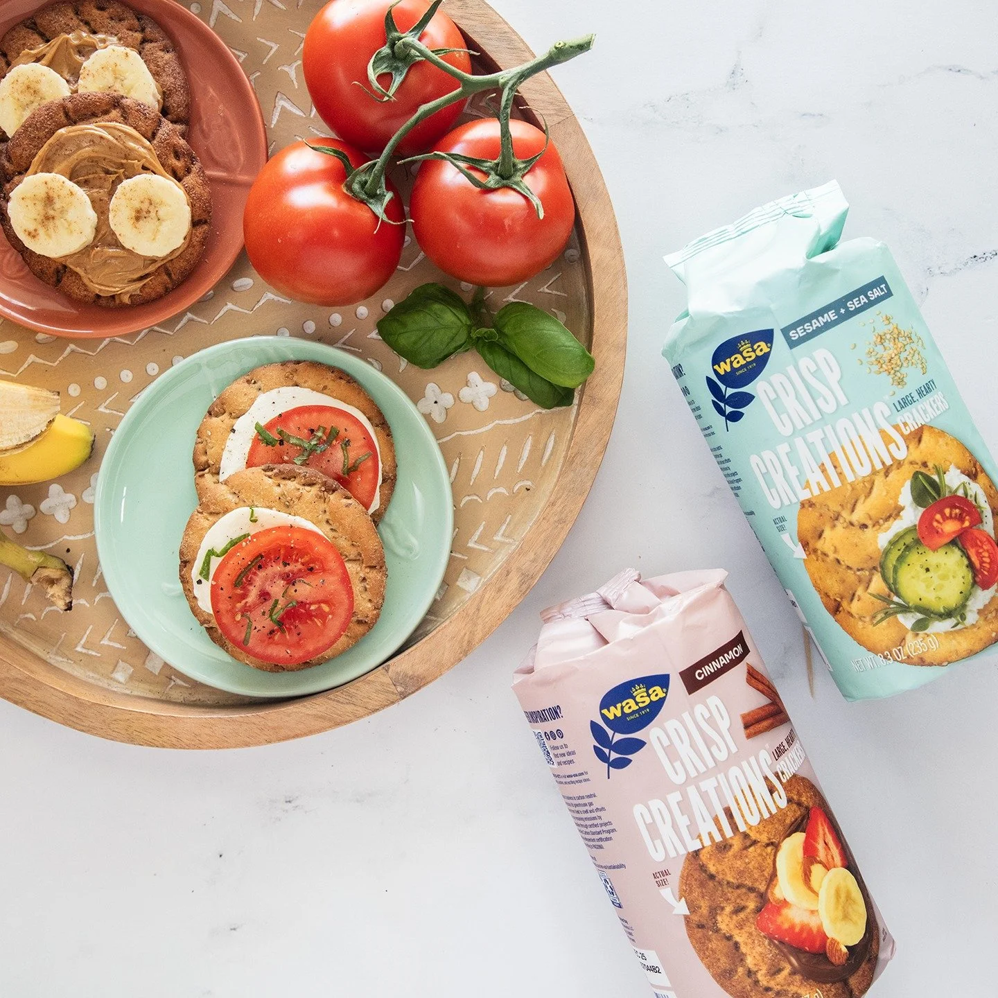 Fresh tomatoes, basil, and packaged crispy crackers on a white surface, with a wooden tray holding two tomato-topped crackers and a small plate with banana and peanut butter snacks.