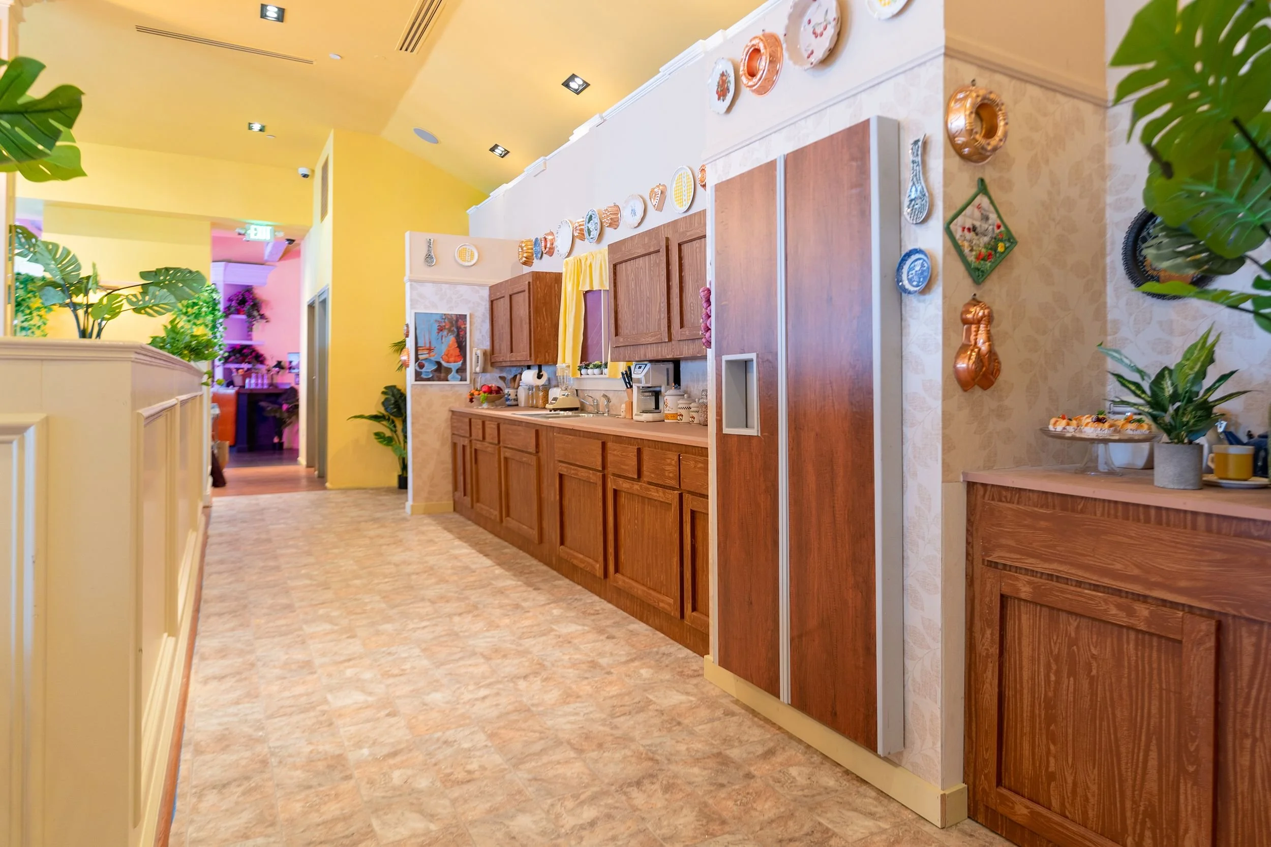 Decorative kitchen area with wooden cabinets, colorful plates on the wall, and tropical plants in a restaurant or cafe setting.