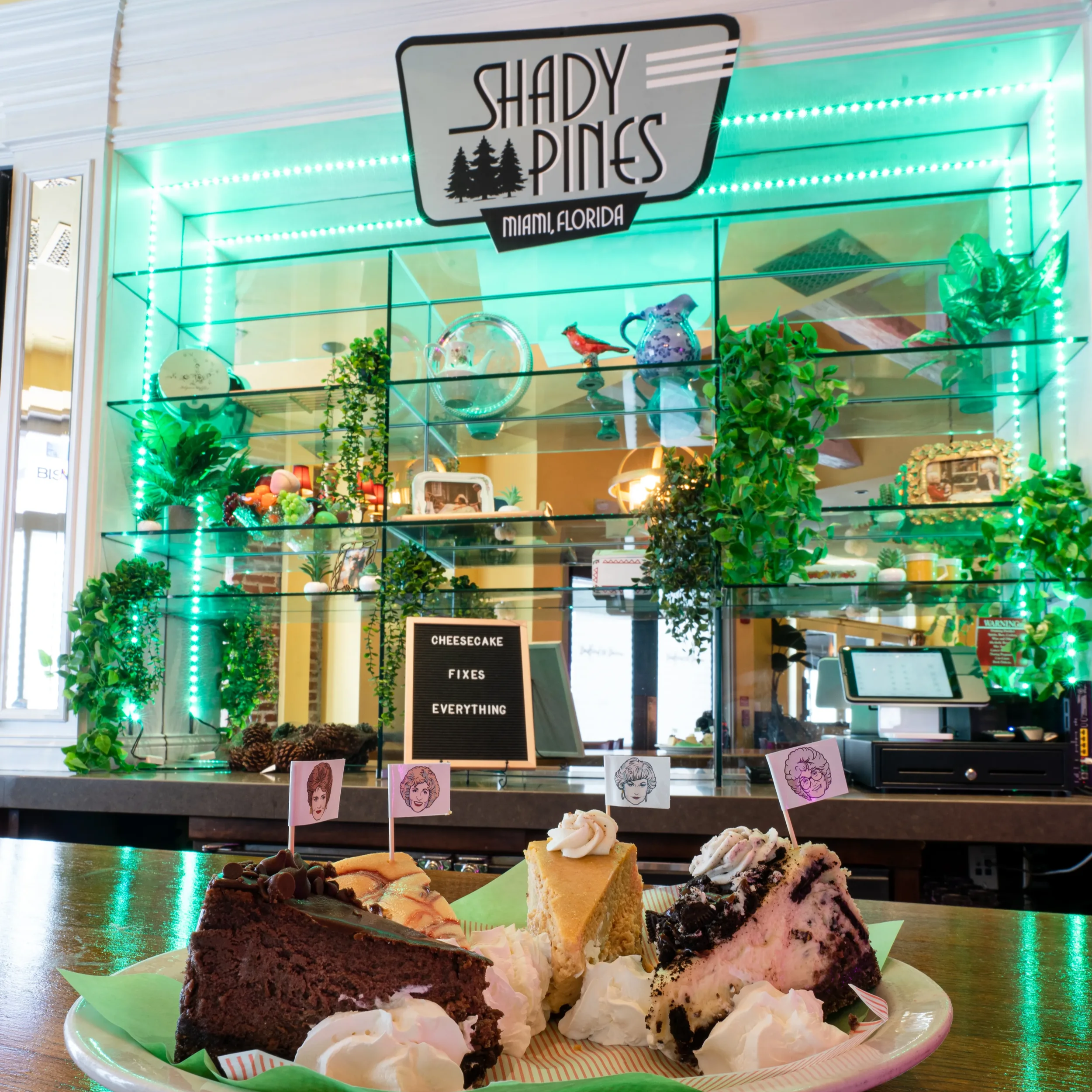 A plate of assorted slices of cheesecake with whipped cream on a wooden table in front of a glass display case filled with plants, glassware, and decorative items in a cafe called Shady Pines, Miami, Florida.