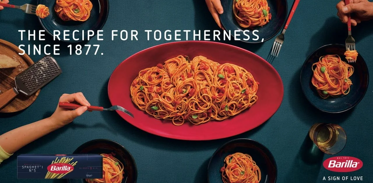 A family dinner table with a large plate of spaghetti and smaller plates of spaghetti, chopsticks, and forks, along with a package of Barilla spaghetti.