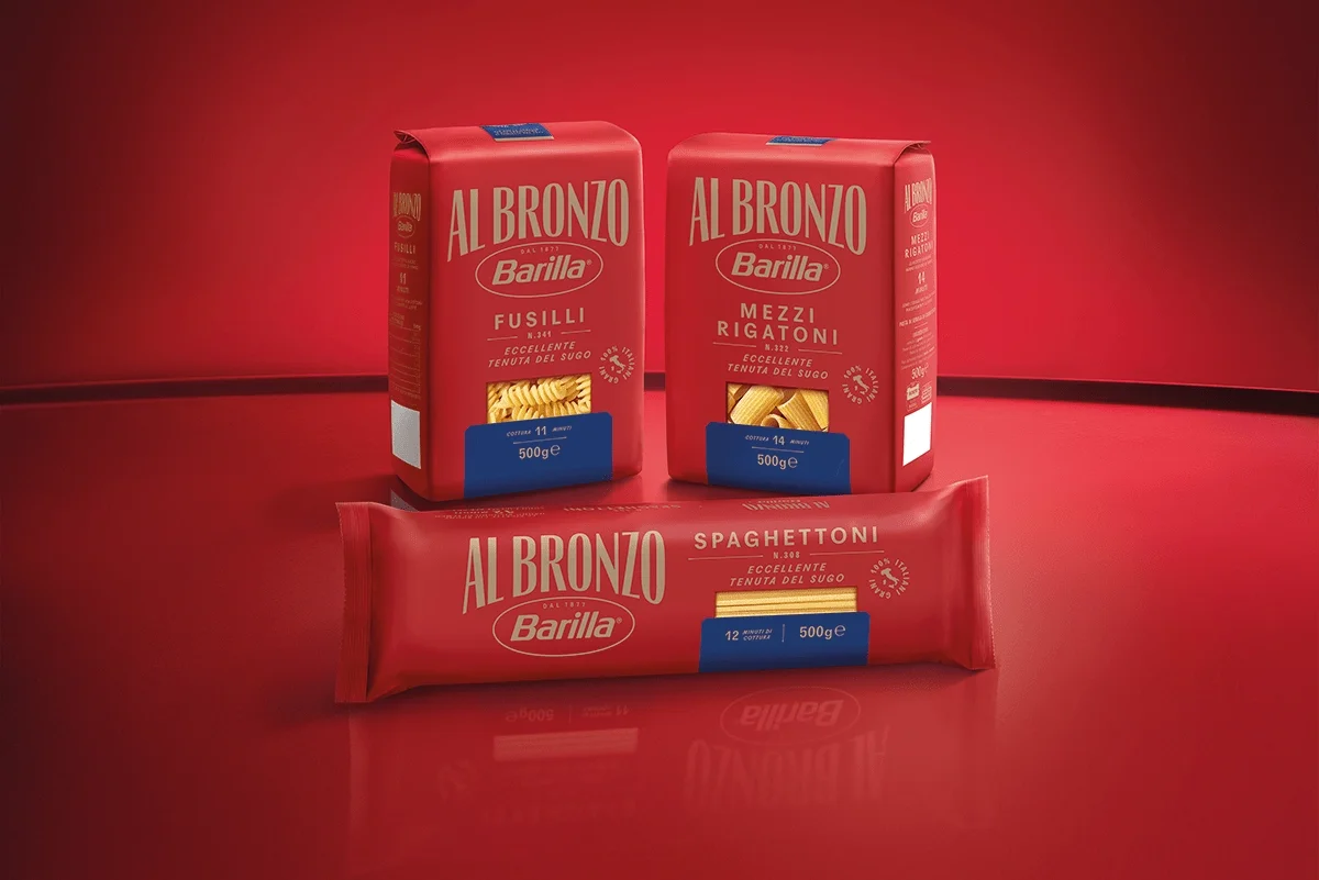 Three packages of Barilla AL BRONZO pasta on a red surface with a red background. Two are upright and one is lying down.