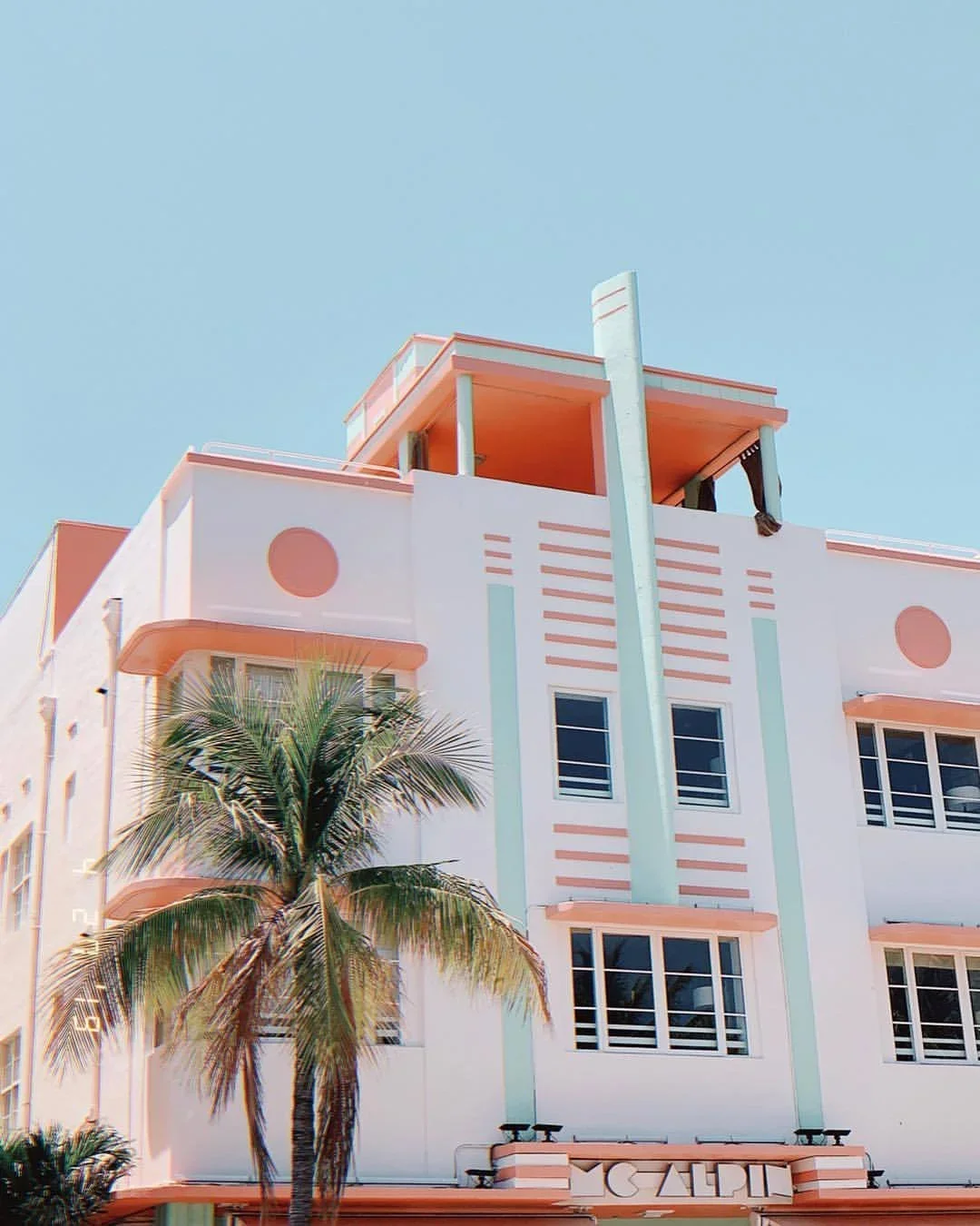 Art Deco style hotel with pastel pink and mint green accents, a tall palm tree in front, and a clear blue sky.