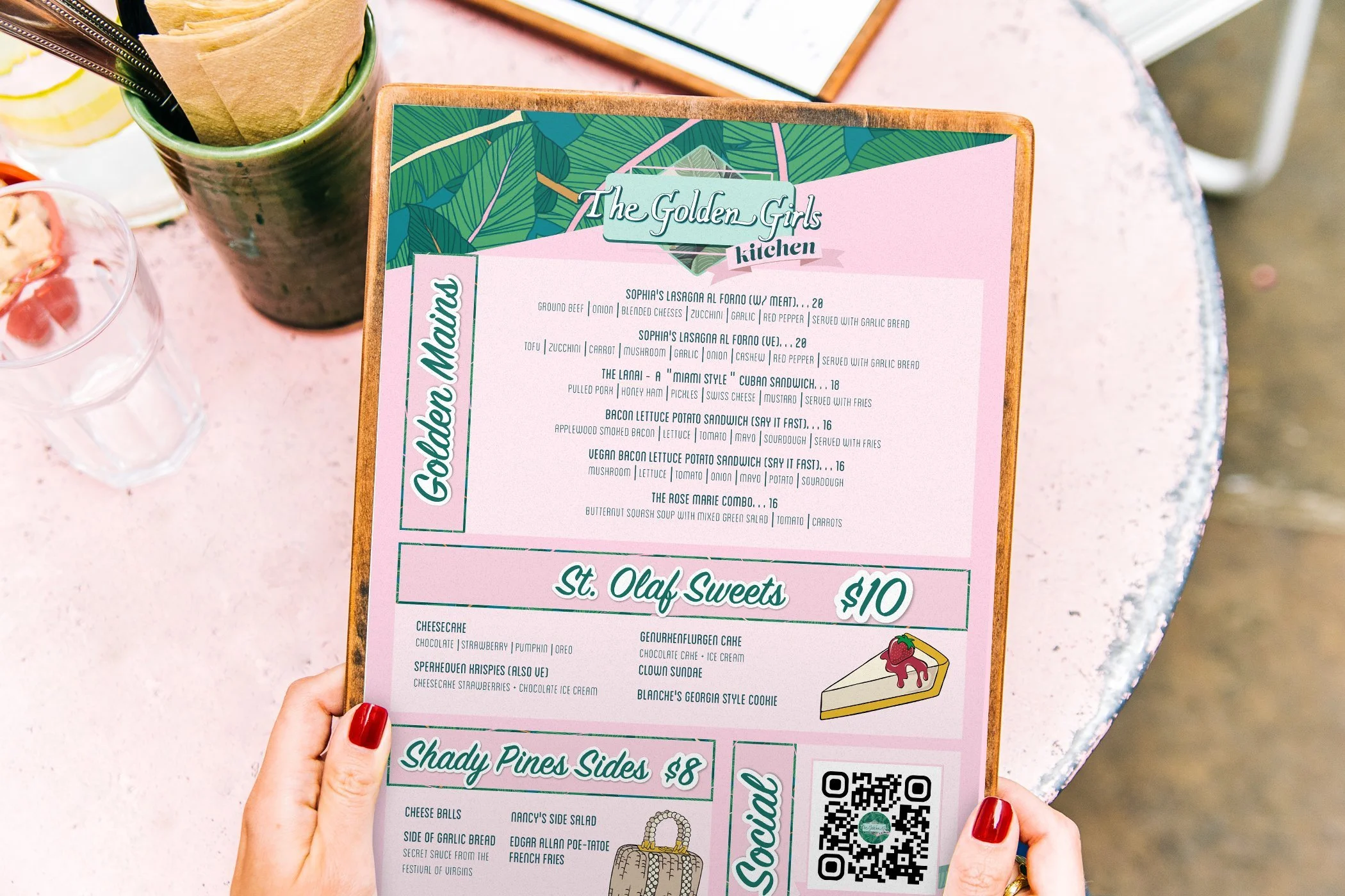 A menu board from The Golden Girls Kitchen on a pink table, displaying food and dessert options.