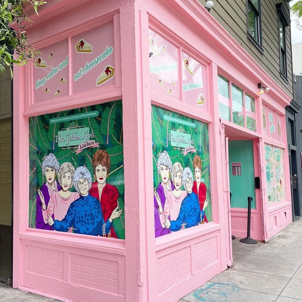 The exterior of a pink-painted building with cartoon illustrations of four women on the window with tropical leaf background. Text on the window says 'The Golden Girls Kitchen' and 'Keep calm and eat cheesecake'.