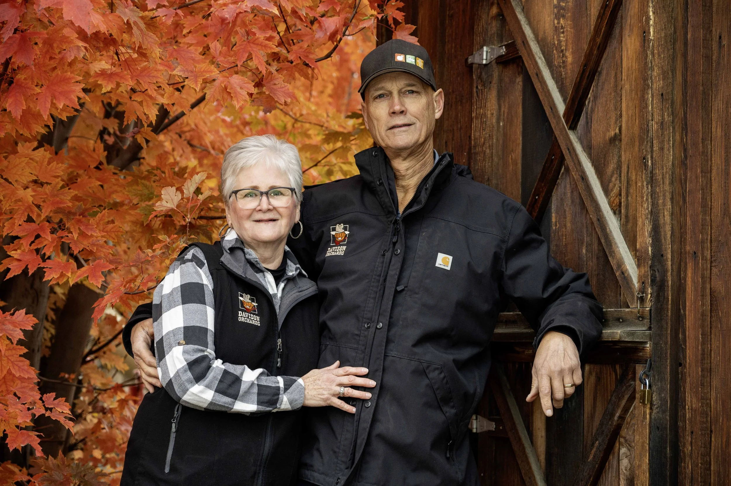 Tamra and Tom Davison standing outdoors in front of a wooden shed with autumn-colored maple leaves in the background
