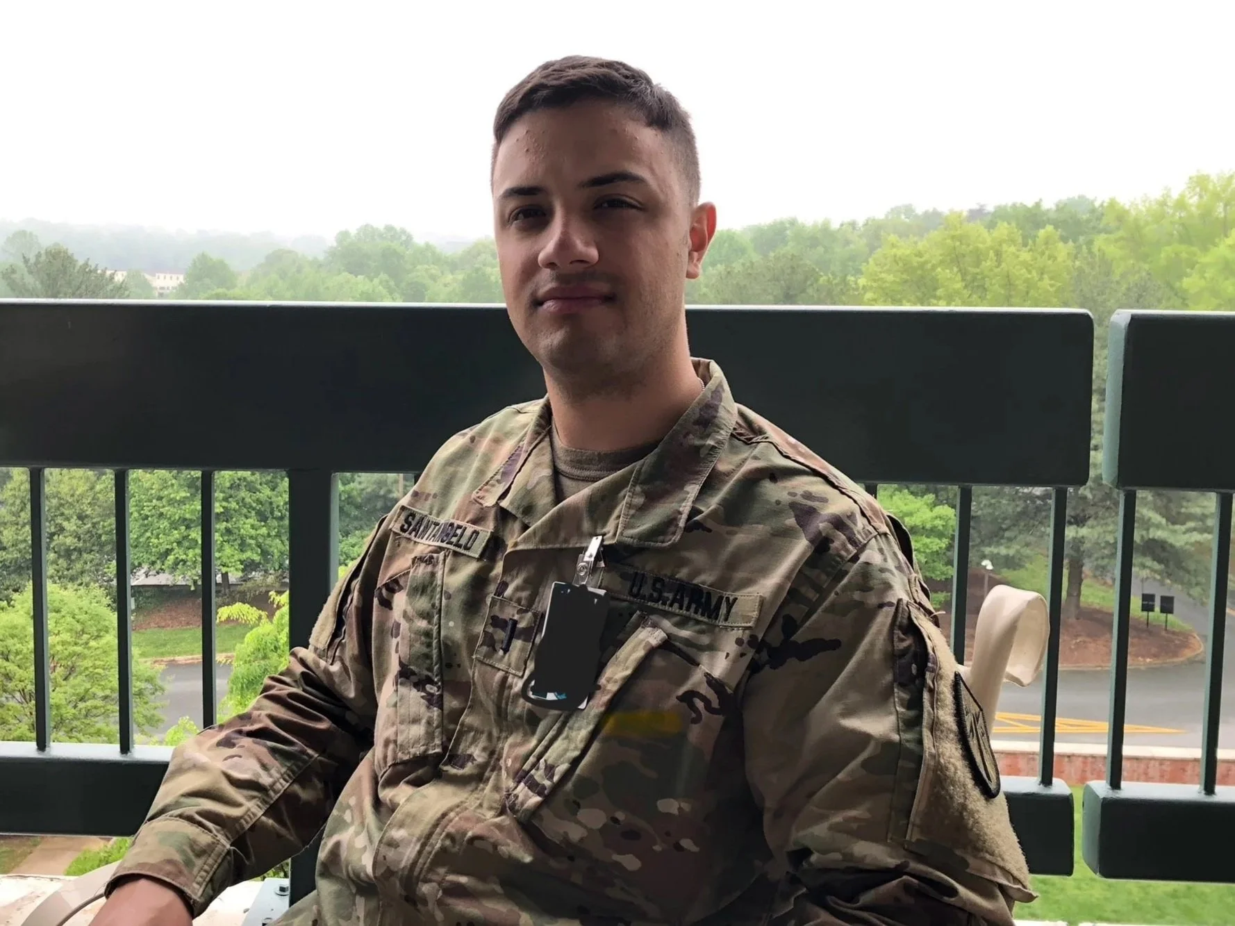 A young man in military camouflage uniform sitting on a balcony with a green, wooded landscape and overcast sky in the background.