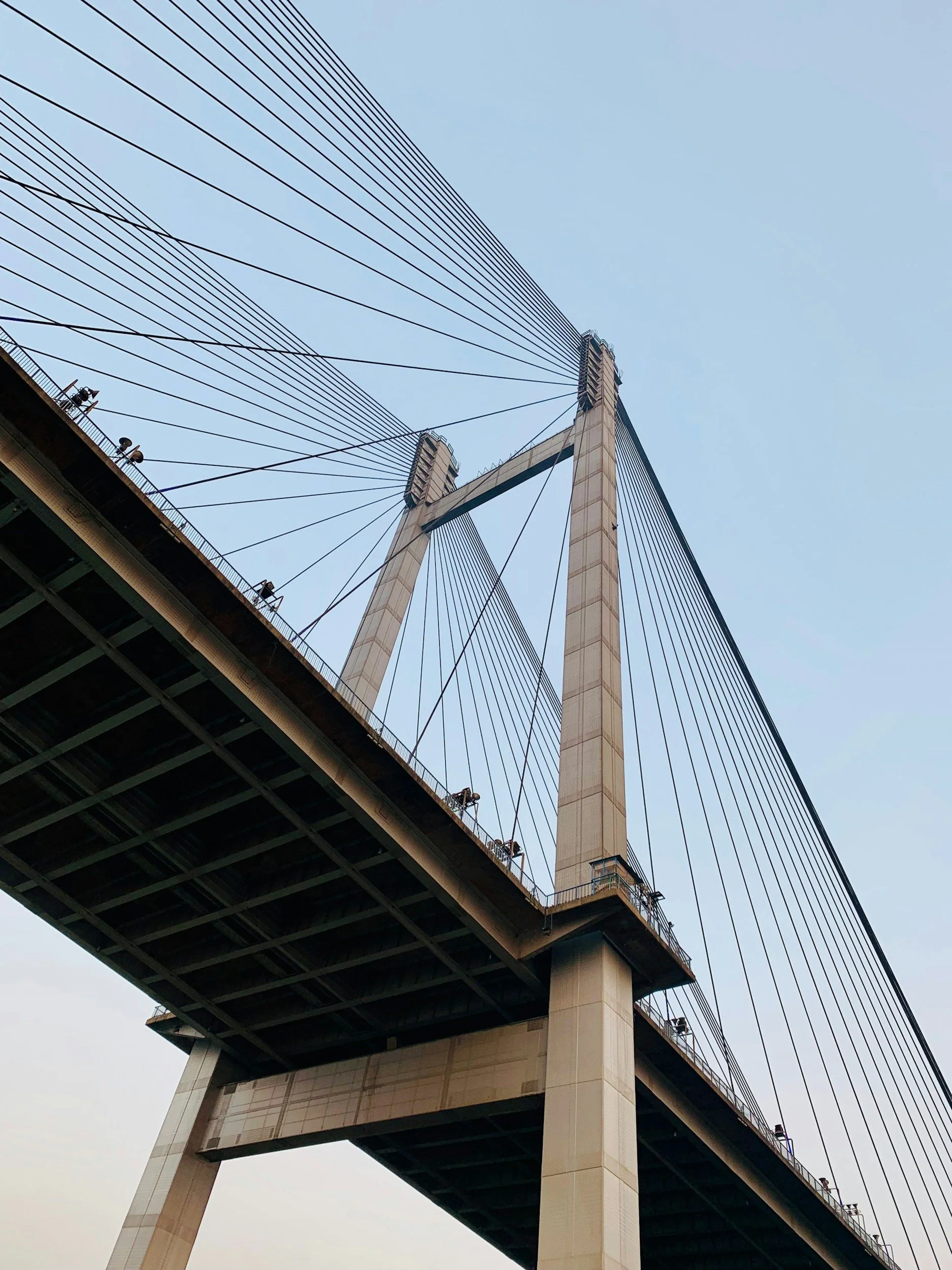 Low-angle view of a large suspension bridge with concrete towers, steel cables, and a roadway beneath a clear sky.