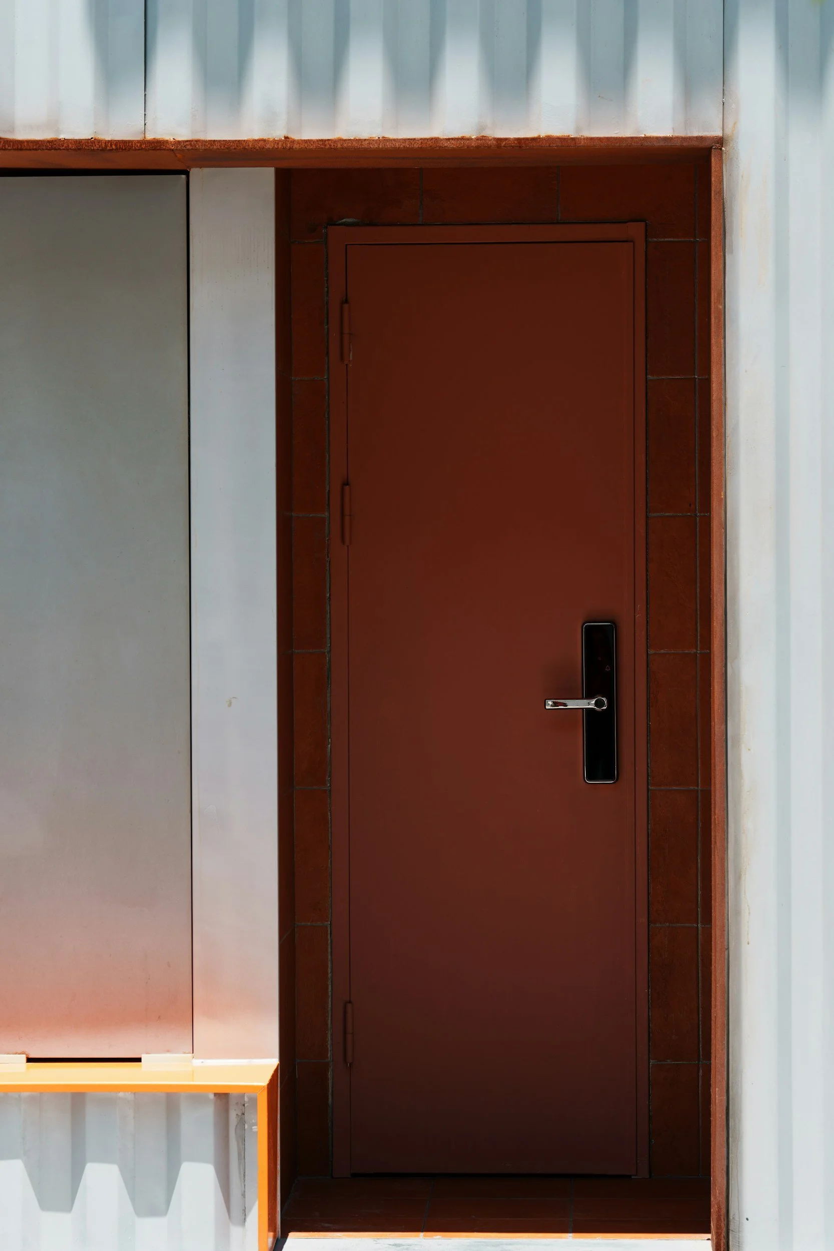 A red metal door with a digital lock system, set within a building with metal siding and brick trim.