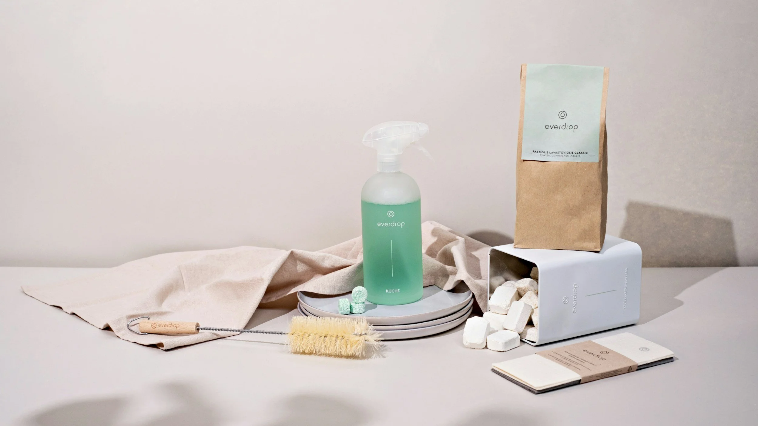 Cleaning supplies including a spray bottle with green liquid, a box of cleaning tablets, a bag of cleaning tablets, a scrubbing brush, and some plates on a table with a beige cloth.