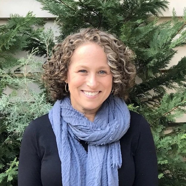 A woman with curly blonde hair smiling outdoors, wearing a black top and a blue scarf in front of greenery.