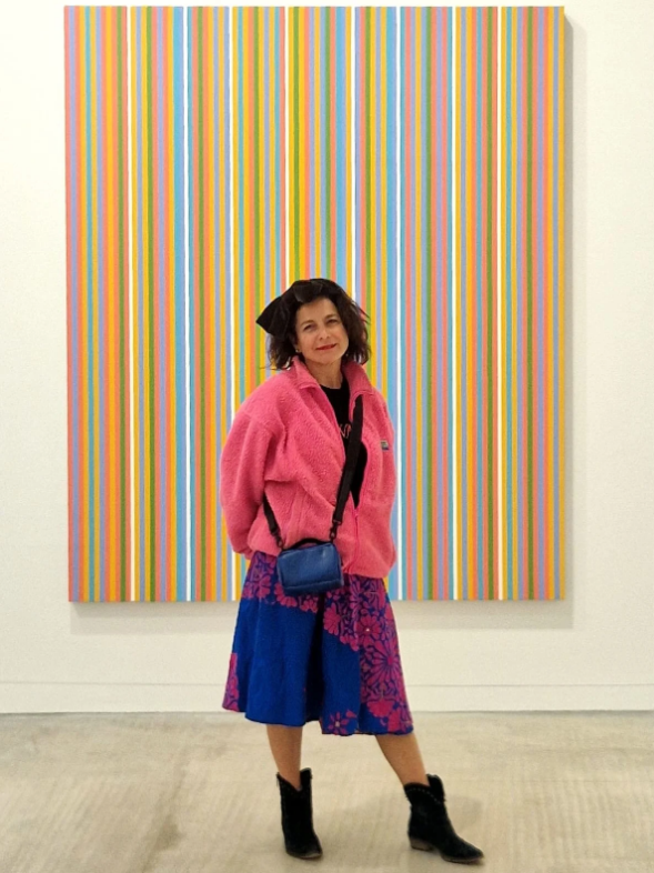 Woman standing in front of an abstract colorful striped artwork, wearing a pink fuzzy jacket, a blue and purple floral skirt, black boots, and carrying a small blue purse.