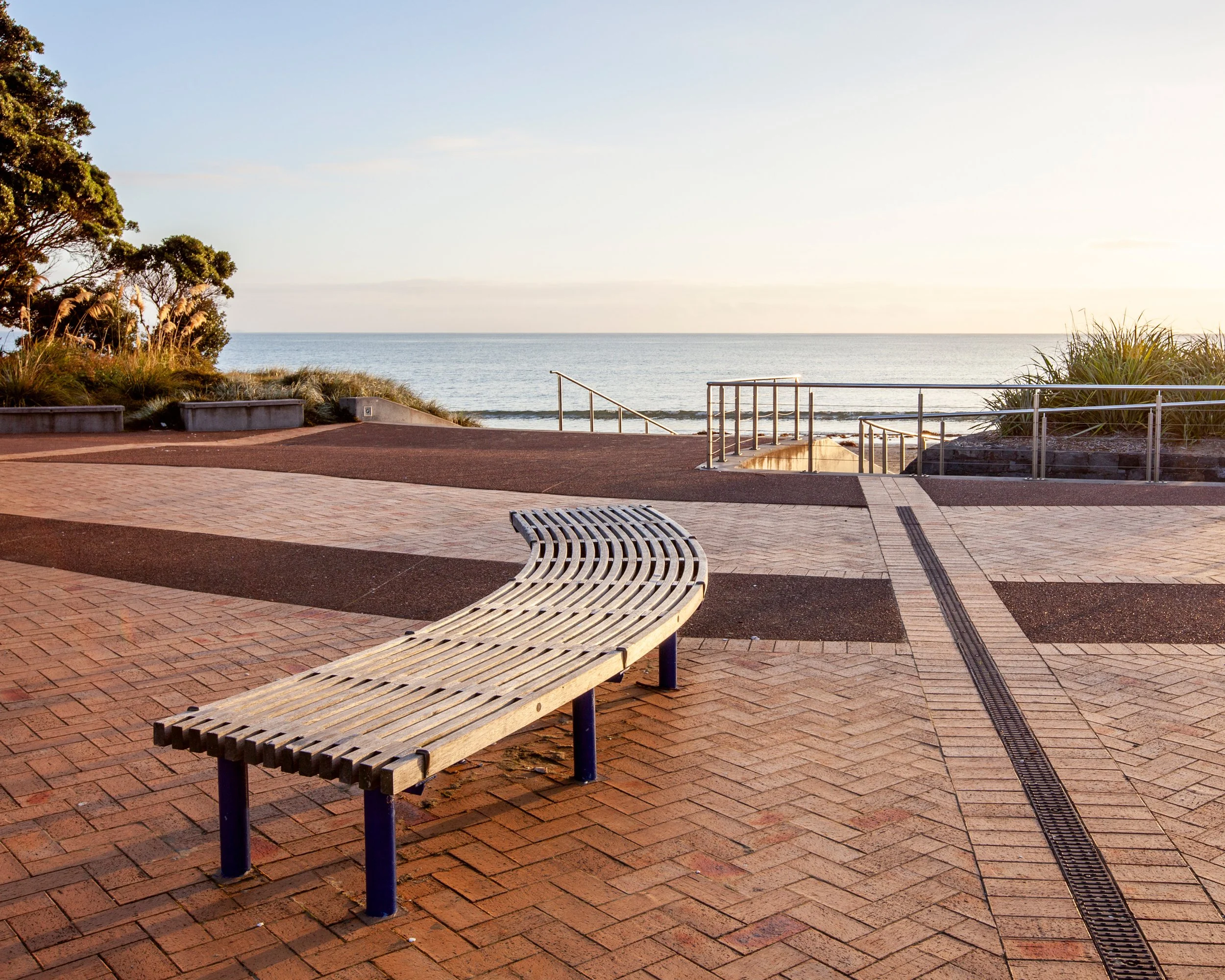 Orewa beachfront promenade with curved bench, brick paving, and beach access at sunrise