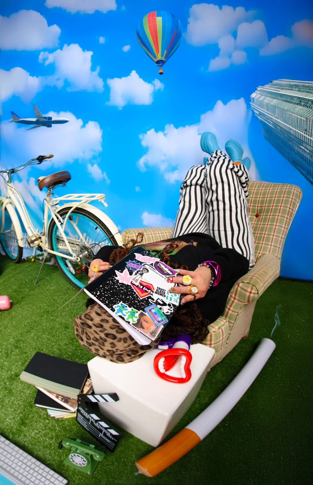 Person lying on a plaid armchair with feet up, surrounded by various items including a comic book, a toy megaphone, a bicycle, a stack of books, and a green rotary phone. The background depicts a blue sky with clouds, a hot air balloon, an airplane, and a tall building, creating a surreal scene.