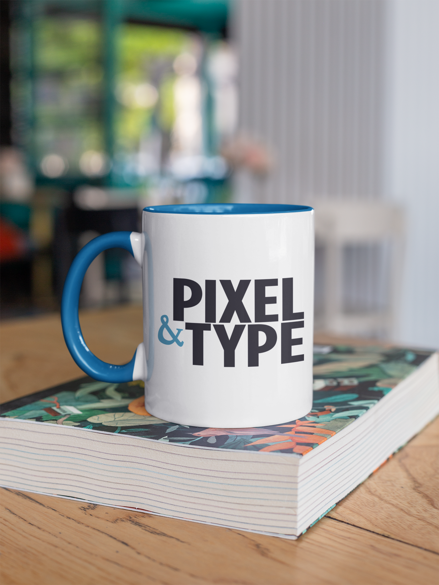 A white ceramic mug with a blue handle and blue interior, with the words 'PIXEL & TYPE' printed on it, sitting on top of an open book with a colorful illustrated cover, on a wooden table.