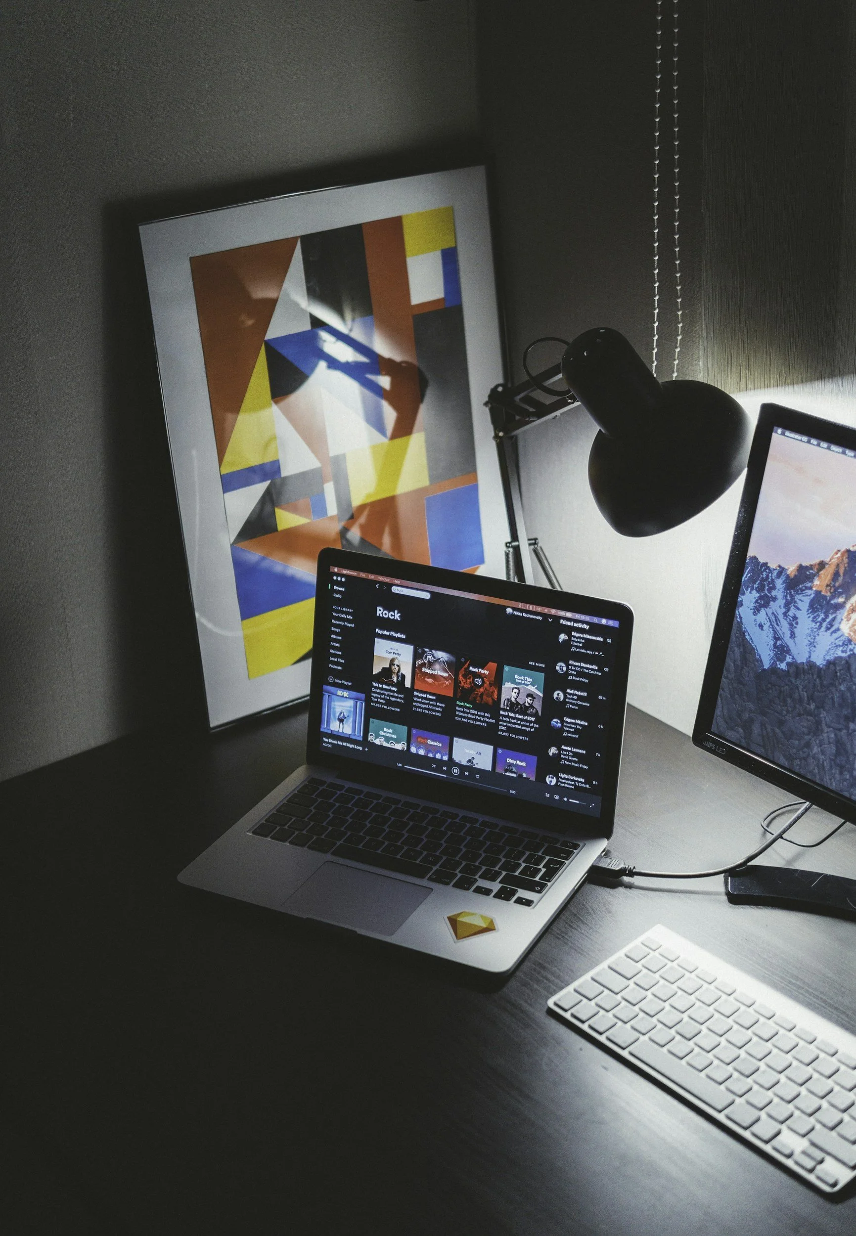 A workspace with a silver MacBook showing a Spotify playlist called 'Rock', a second monitor displaying a mountain landscape, a black desk lamp, a white keyboard, and a colorful abstract framed artwork on the wall.