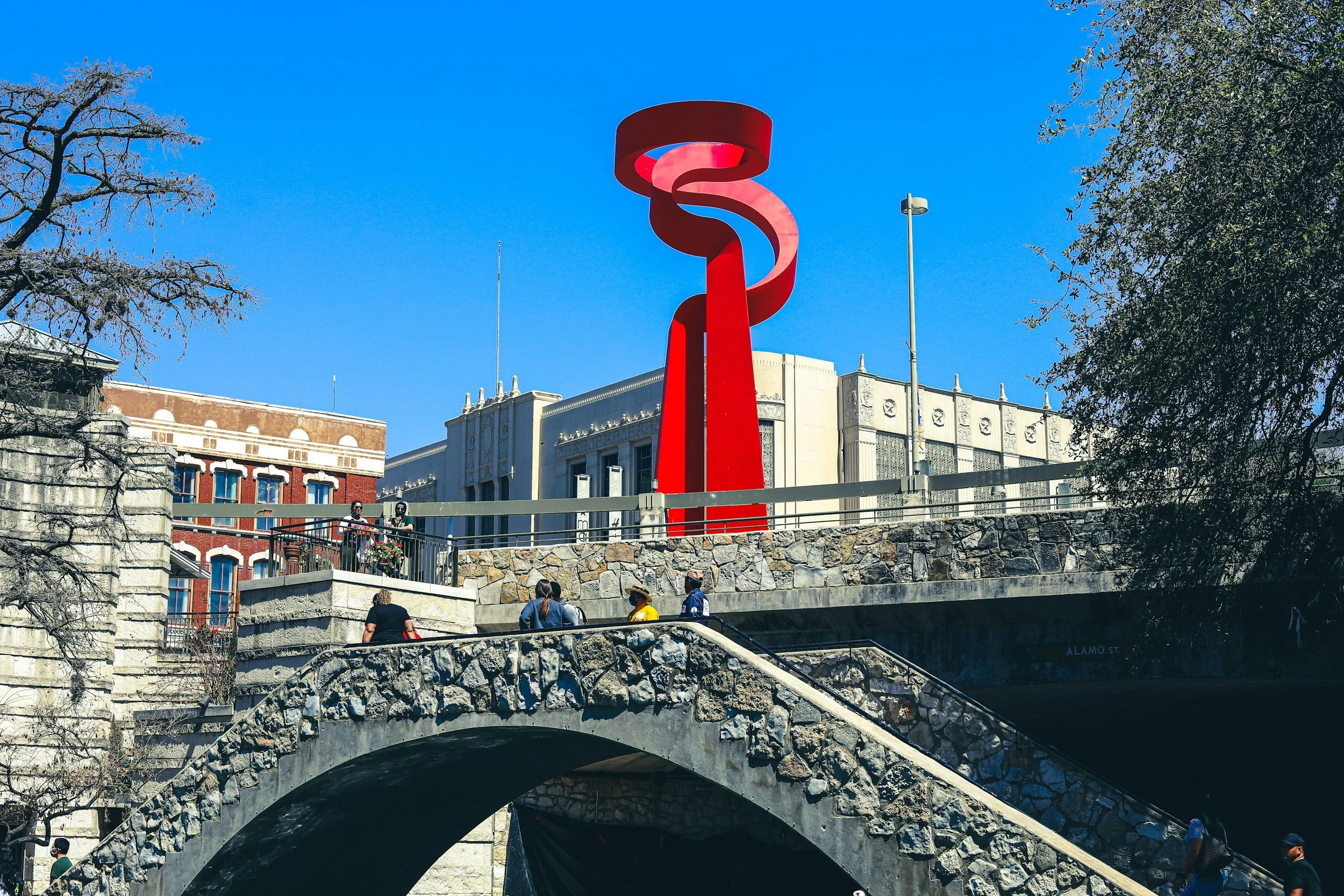 Red abstract sculpture on a bright blue sky background, with buildings and people in the foreground.