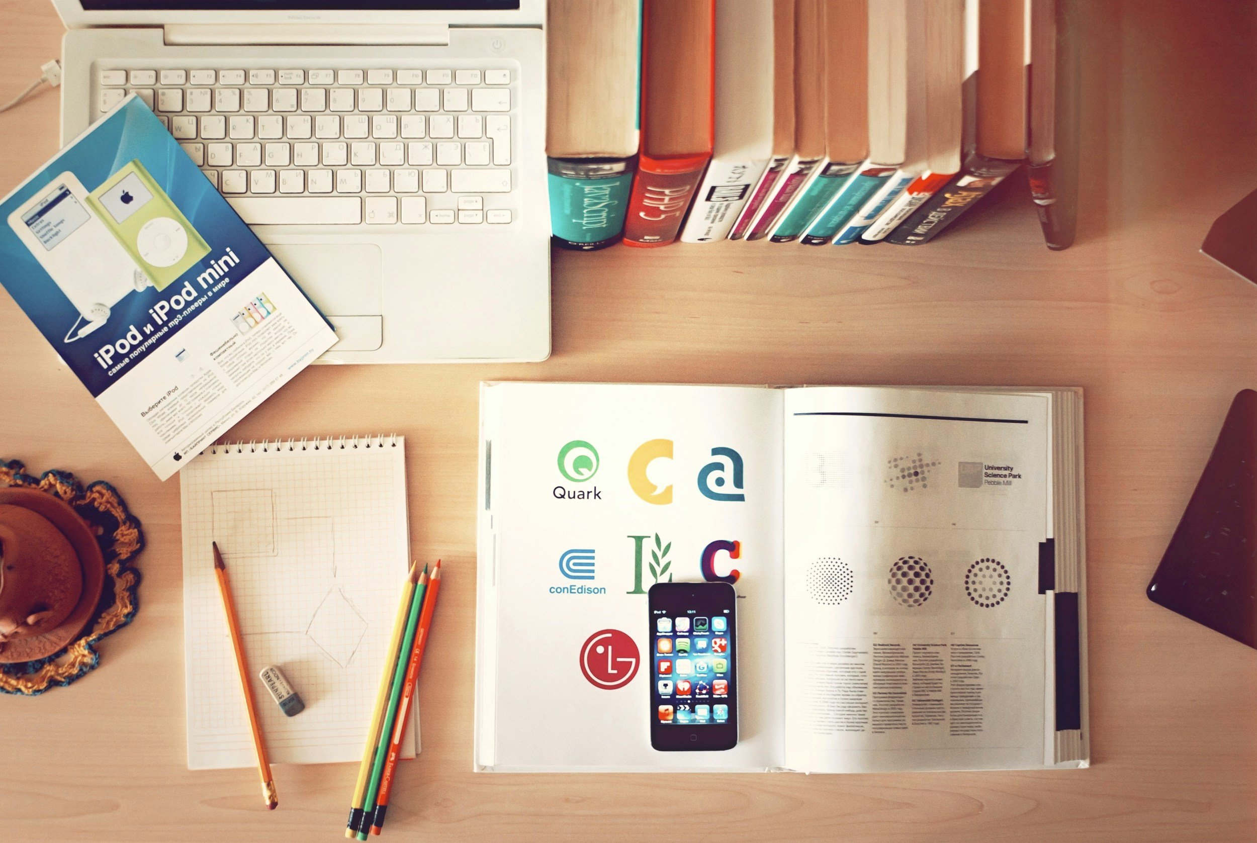 Desk with a white laptop, colorful pencils, a notepad, a smartphone with app icons, books, a brochure about iPod and iPhone, an open book with logos and text, and a decorative hat on the left side.