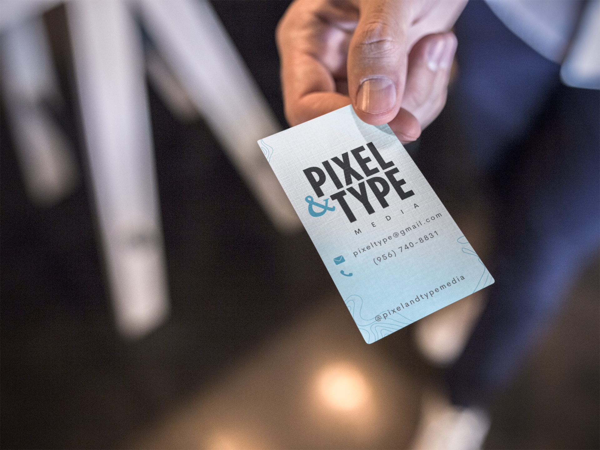 Close-up of a person's hand holding a business card with the text 'PIXEL & TYPE MEDIA' and contact information. The background is blurred.