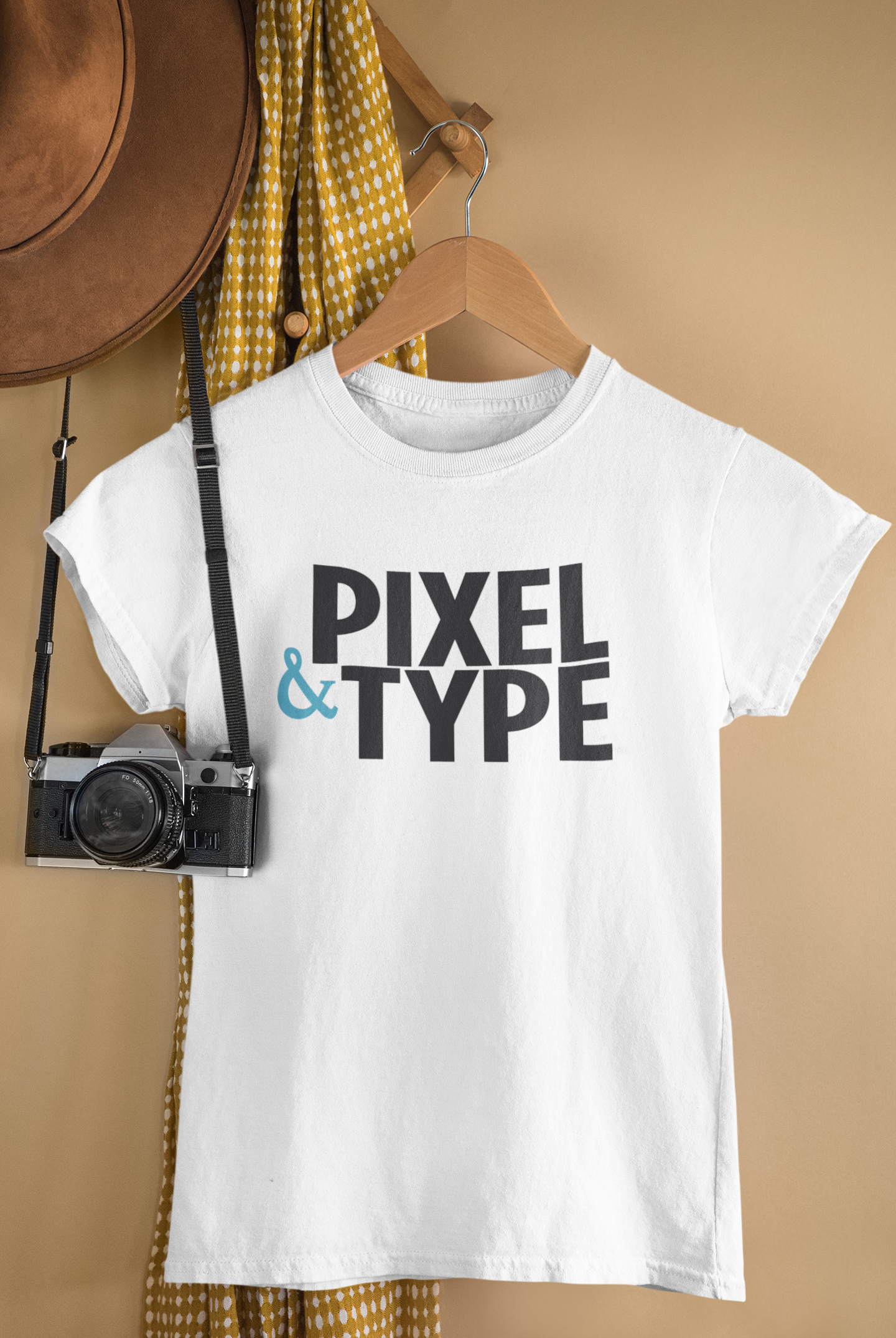 A white t-shirt with the text 'PIXEL & TYPE' printed on it hanging on a wooden hanger. To the left, there is a vintage camera hanging from a strap. In the background, a yellow polka dot dress and a brown wide-brimmed hat are hanging on a wooden rack against a beige wall.
