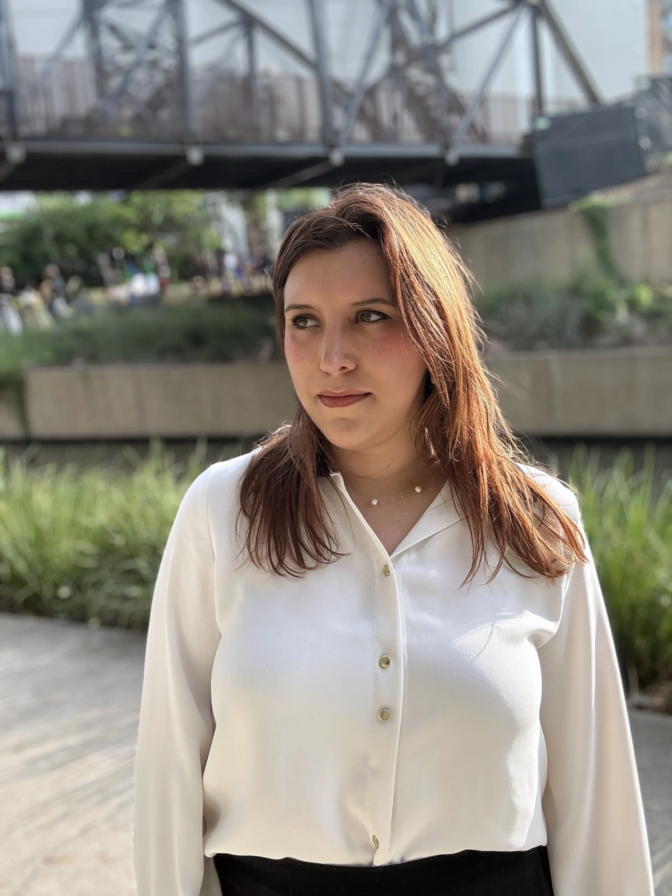 A woman with reddish-brown hair wearing a white blouse with buttons and a pearl necklace, standing outdoors near a river and bridge in the late afternoon.