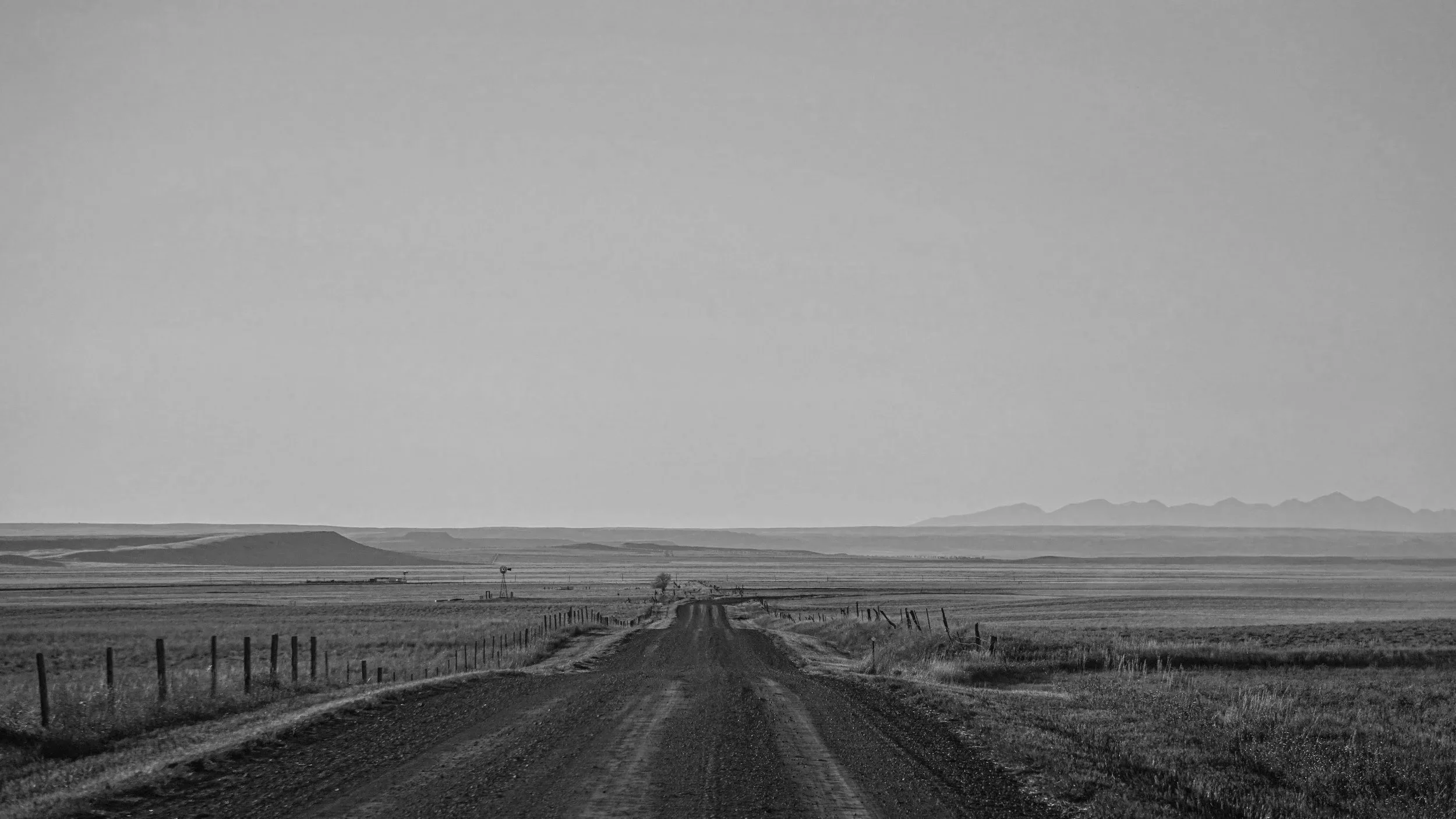 A black and white photo of a rural dirt road stretching into the horizon, flanked by open fields with fences on either side, and distant mountains in the background.