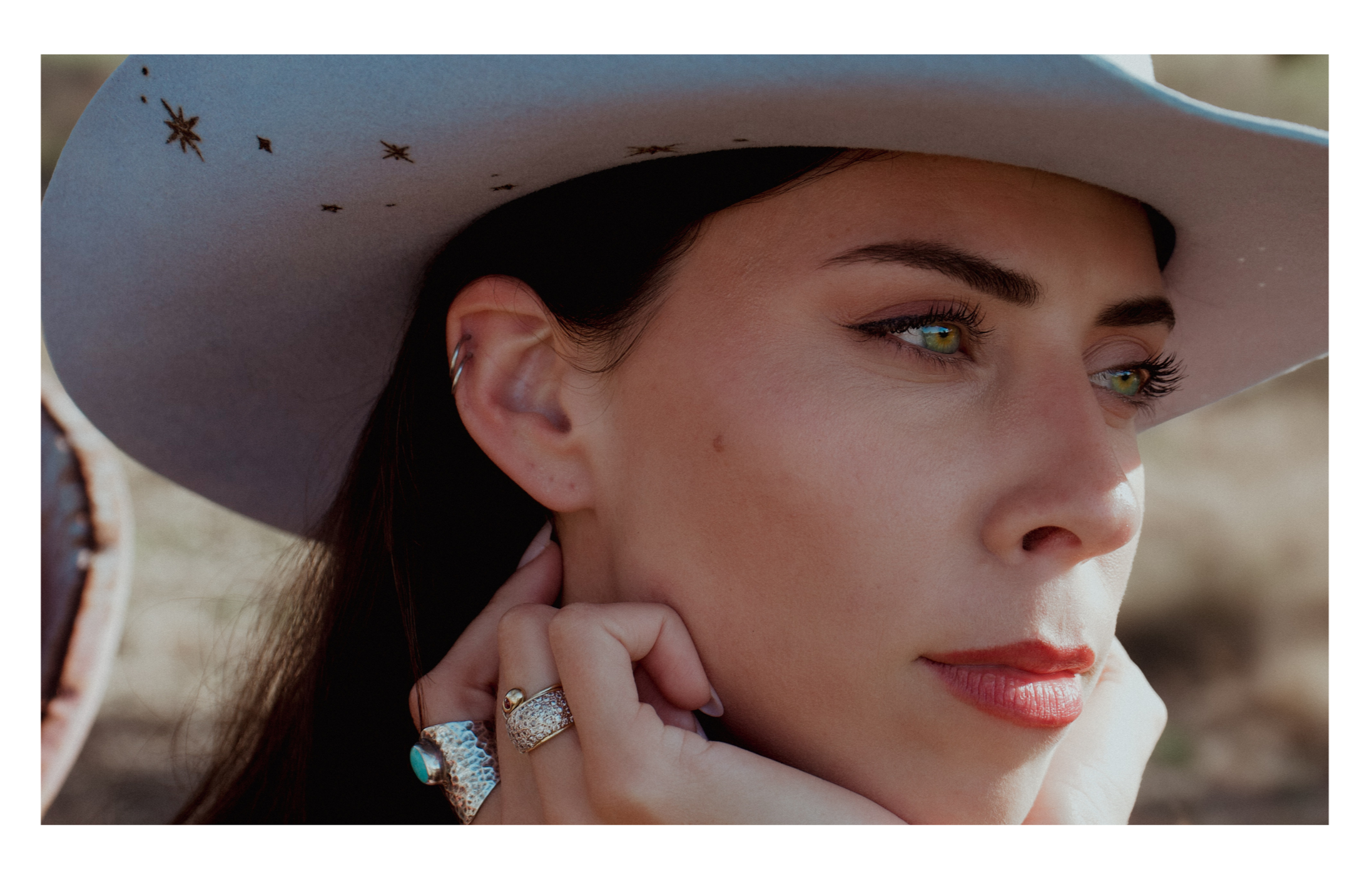 Close-up of a woman wearing a wide-brimmed hat with star and dot patterns, resting her hand on her face, showing jewelry and natural makeup.