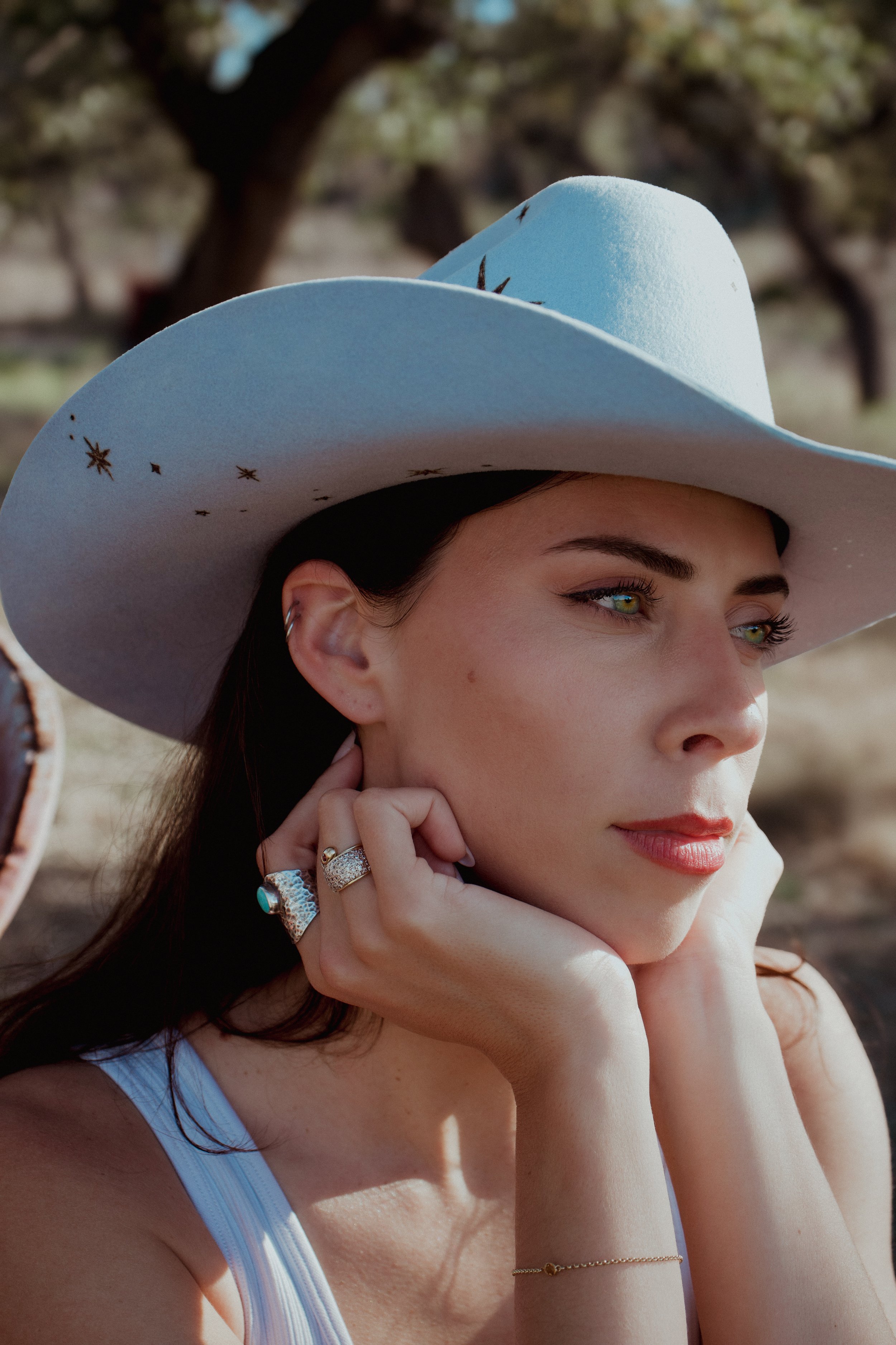 Close-up of a woman wearing a large white cowboy hat, resting her face on her hands outdoors.