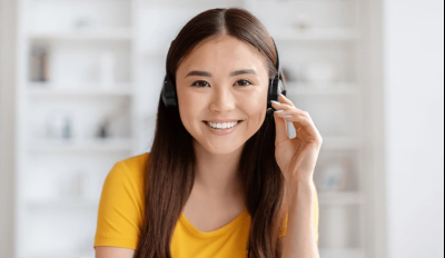 A young woman with long dark hair, wearing a yellow top, smiling and talking on a headset in a bright, modern room.