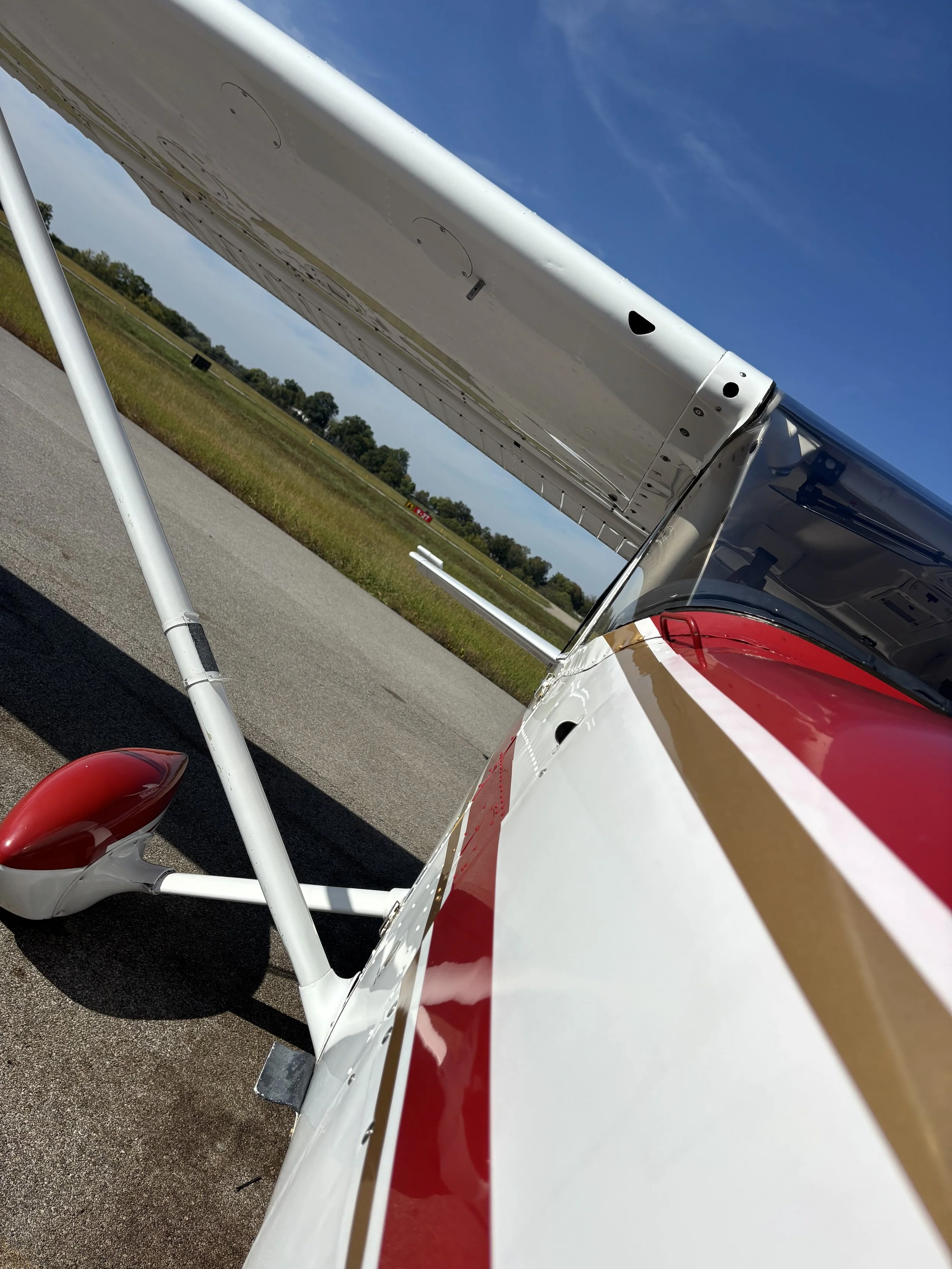 Close-up of a kit plane on the tarmac, showing the white wing with red and gold stripes, and the tail wheel with a red cover, under a blue sky.