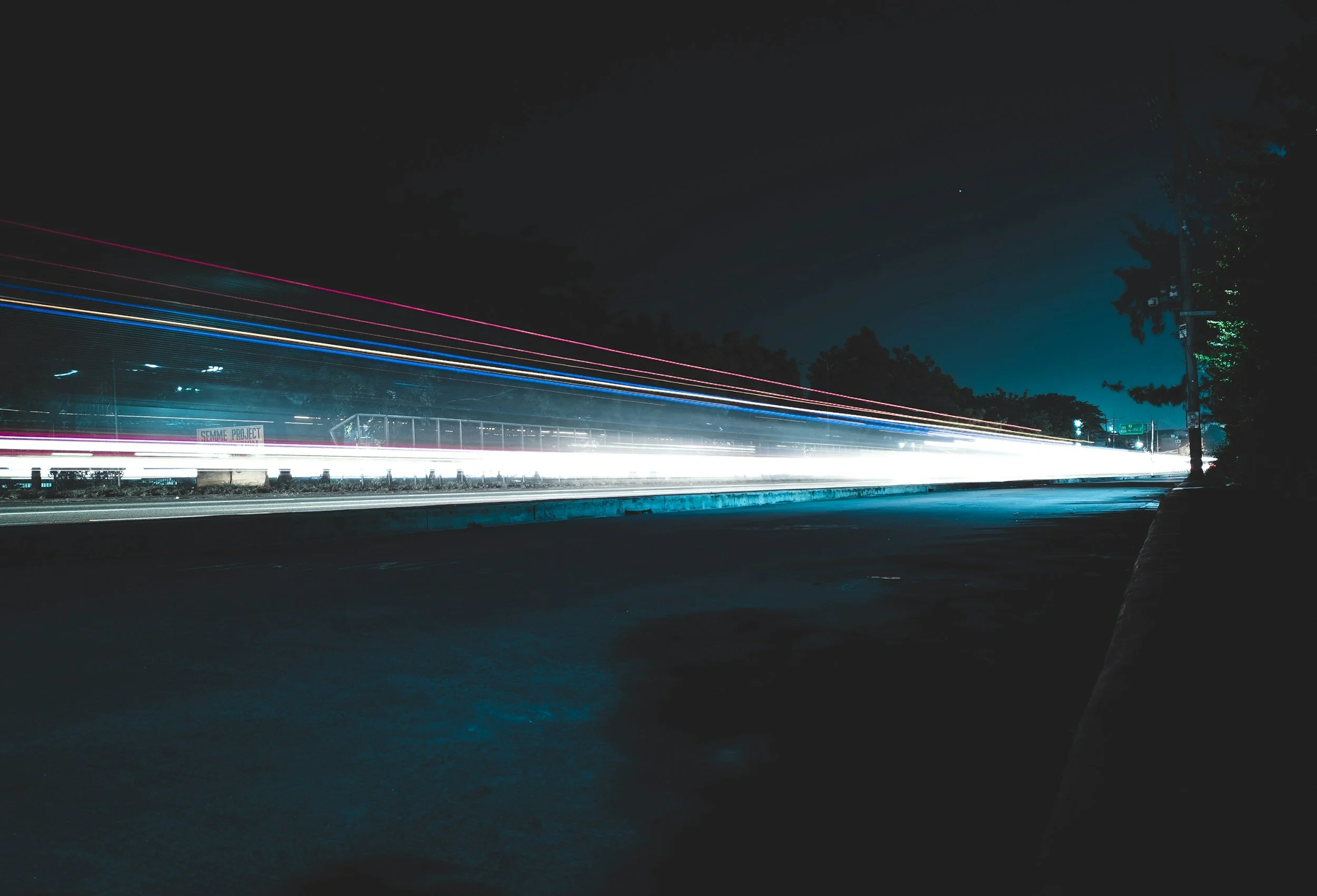 Nighttime scene of a highway with streaks of light from moving vehicles, and silhouettes of trees against a dark sky.