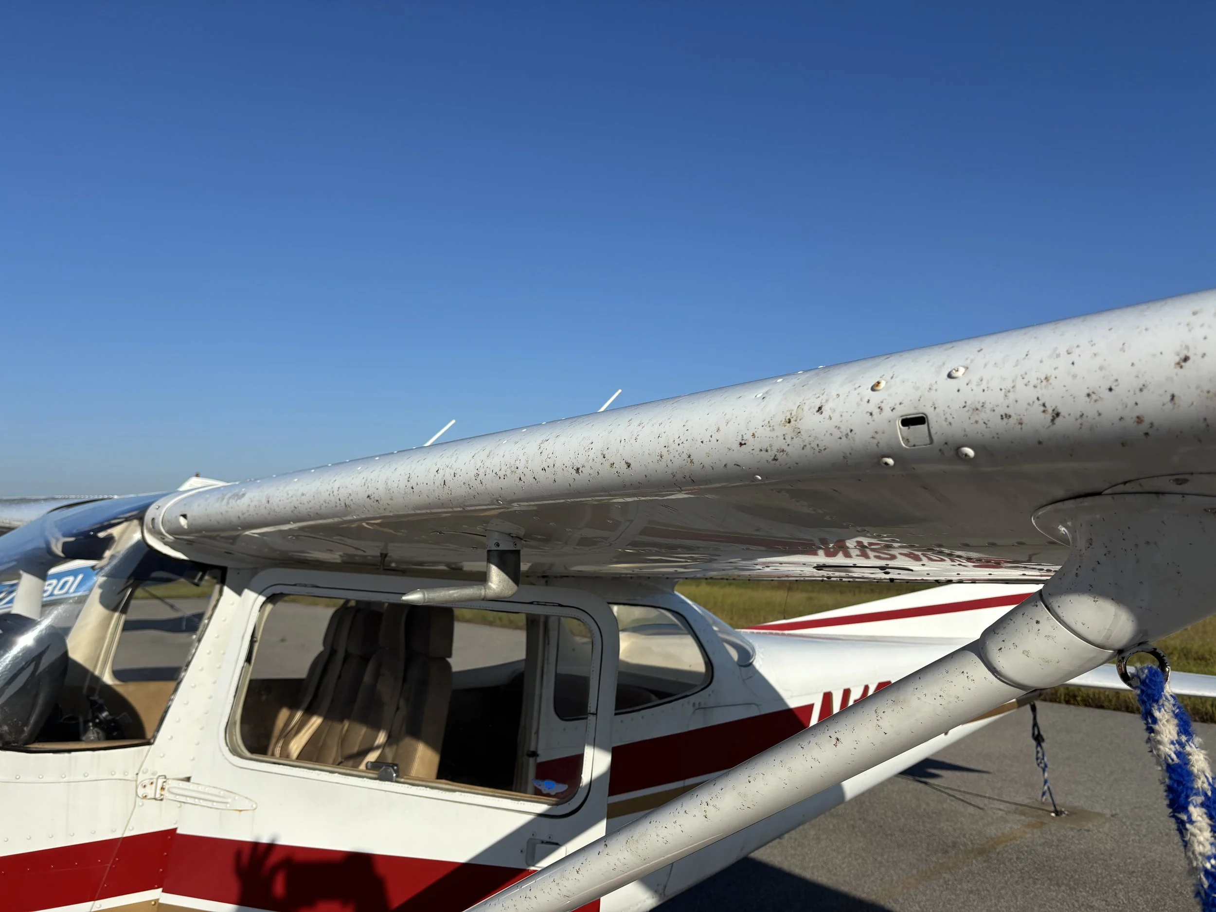 Small white and red airplane parked on a runway under a clear blue sky, with visible dirt and rust on the wing surface.
