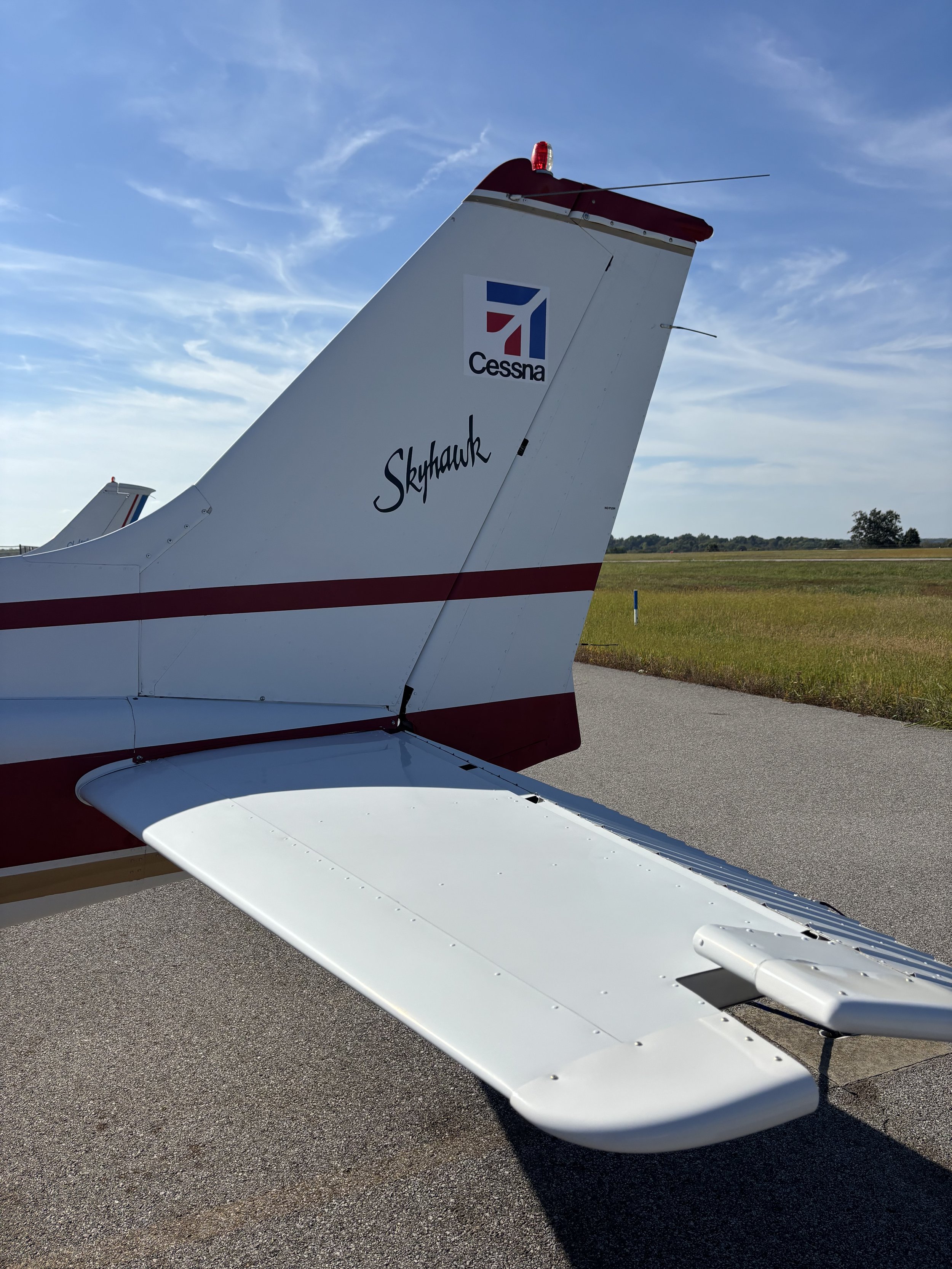 The tail section of a white Cessna Skyhawk airplane with red and blue markings, parked on a tarmac under a partly cloudy sky.