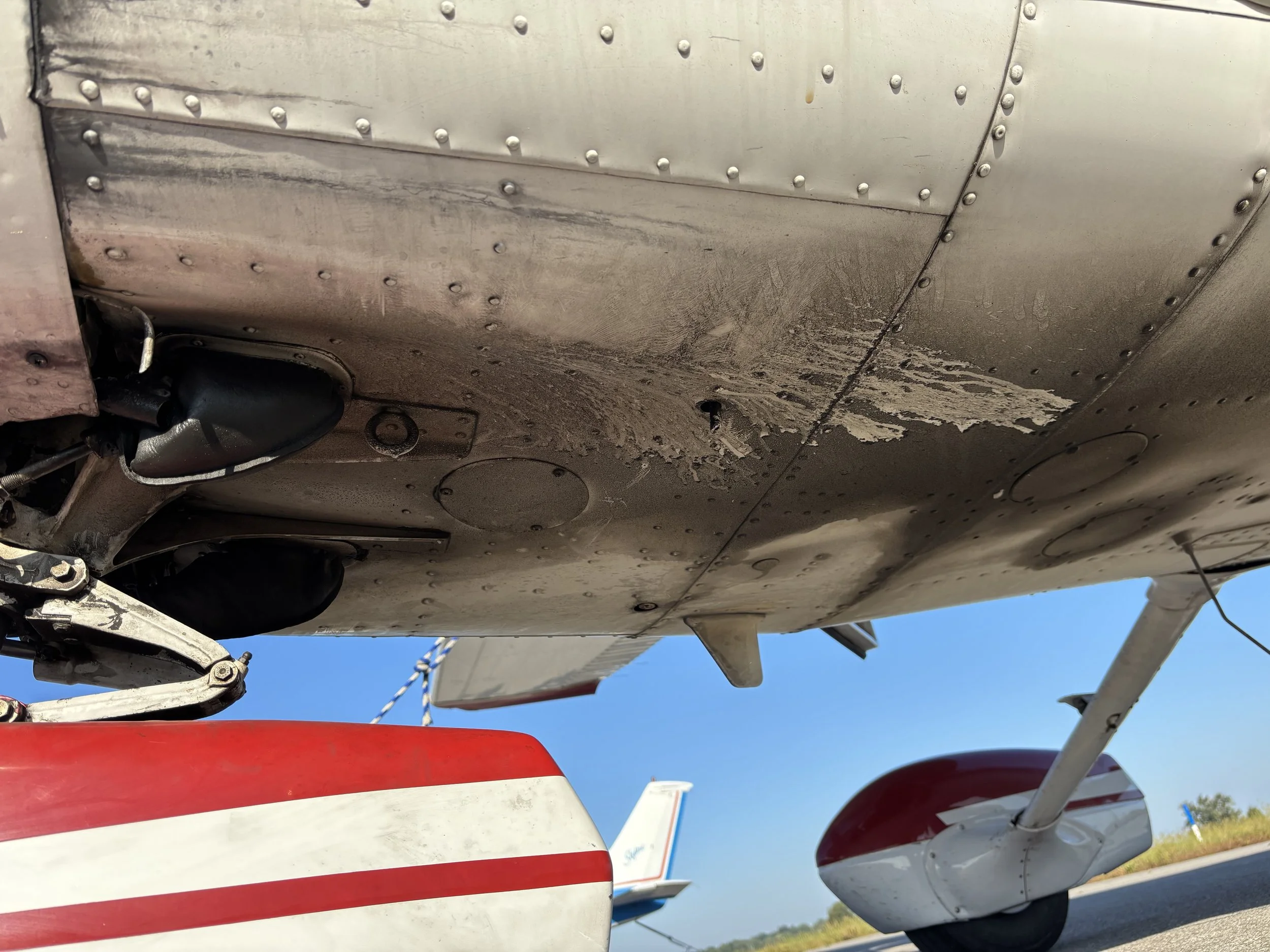 Underside of a small aircraft showing landing gear, fuselage, and part of the wing with a blue sky in the background.
