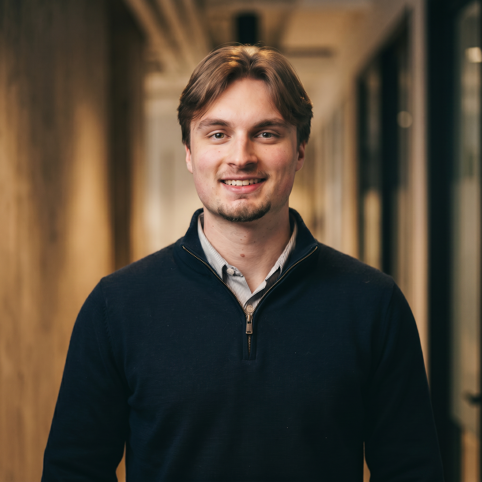 A young man with light brown hair, wearing a dark blue zip-up sweater over a collared shirt, standing in a corridor with wooden-paneled walls, smiling at the camera.