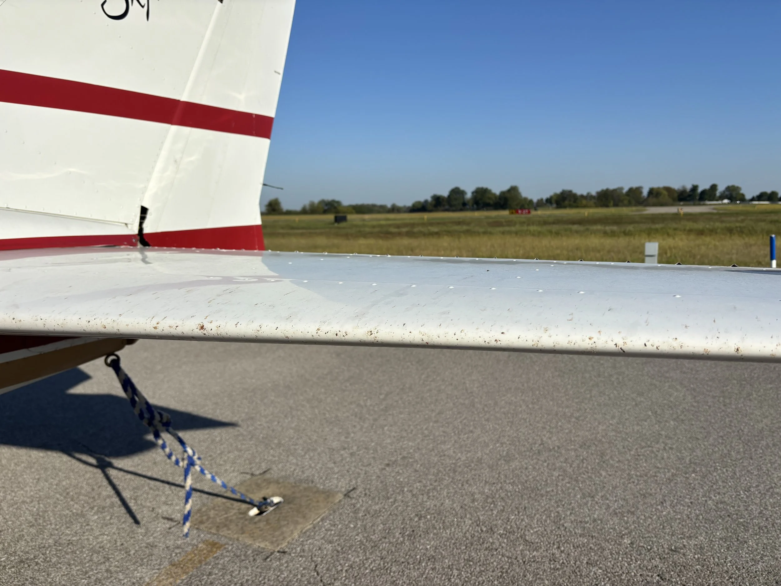 Close-up of a small aircraft's wing and tail section on runway, with grassy field and blue sky in the background.