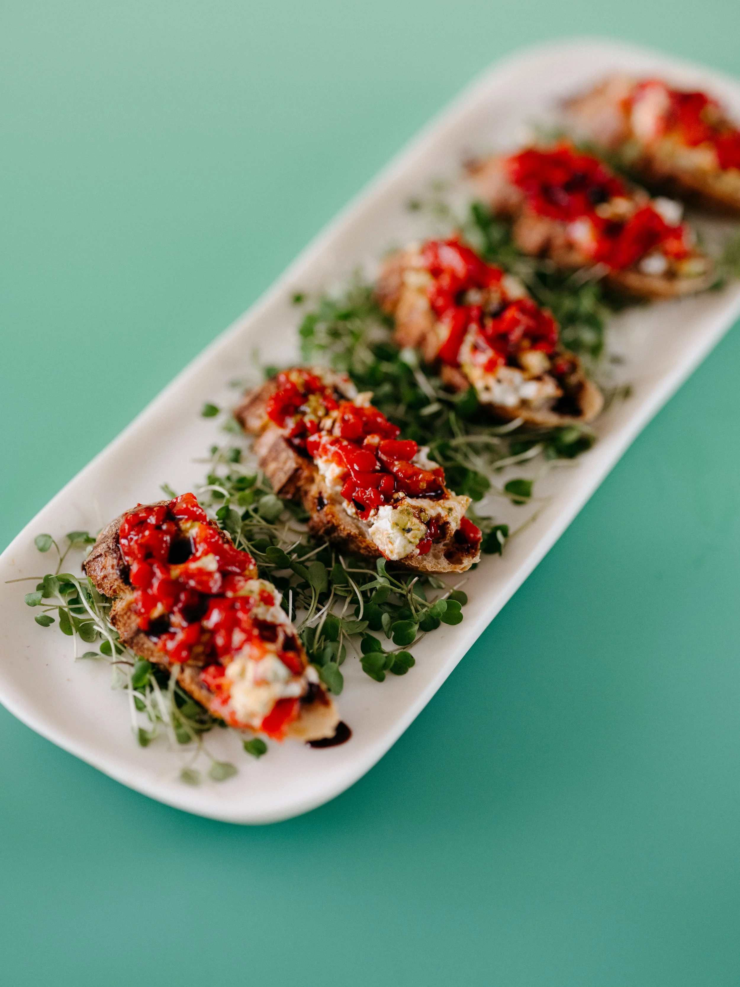 Six pieces of bruschetta topped with tomatoes and cheese, garnished with microgreens on a white platter.