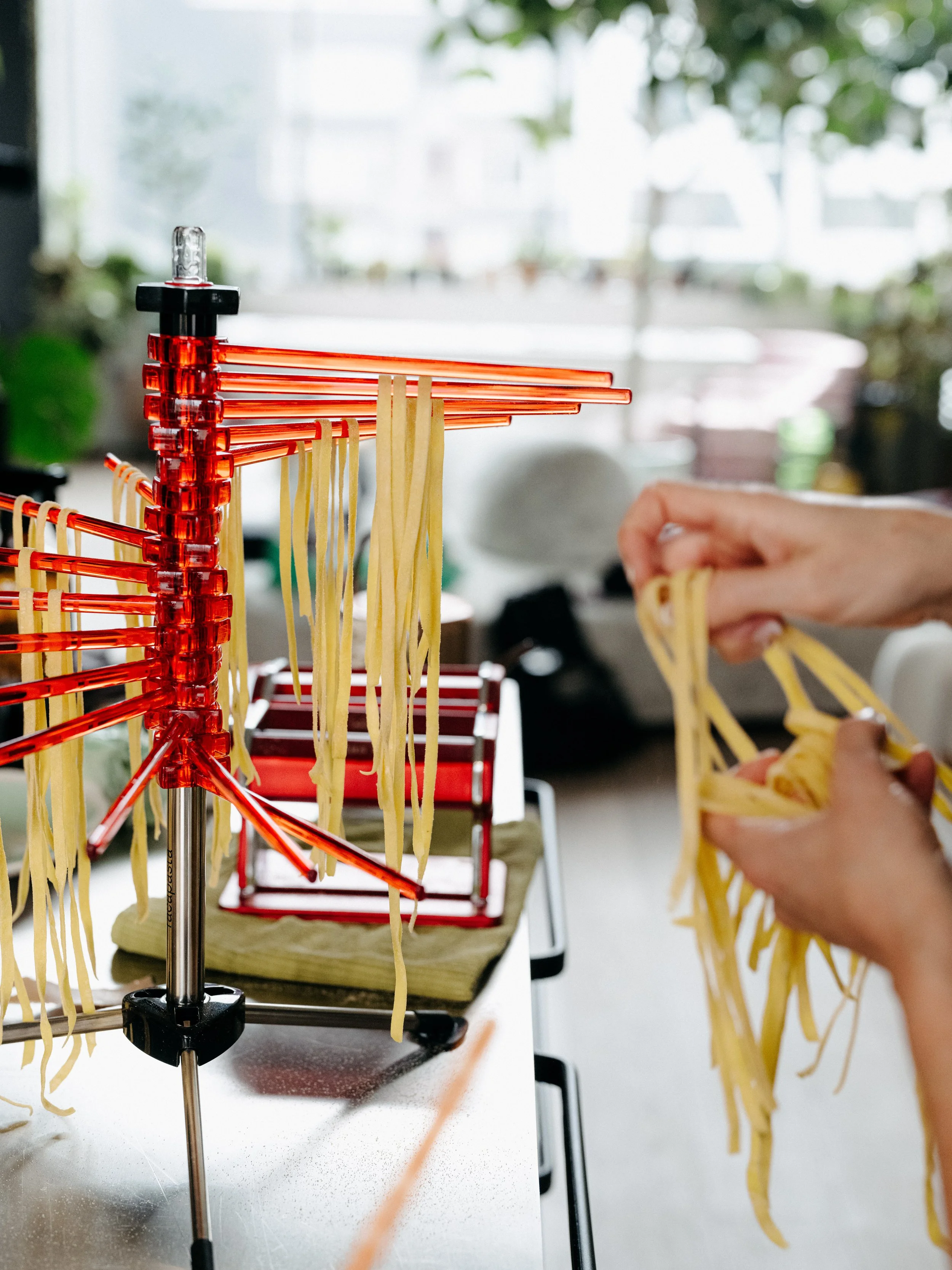 Close-up of a pasta drying rack with freshly made pasta hanging to dry. A person is also seen hand-hanging pasta.