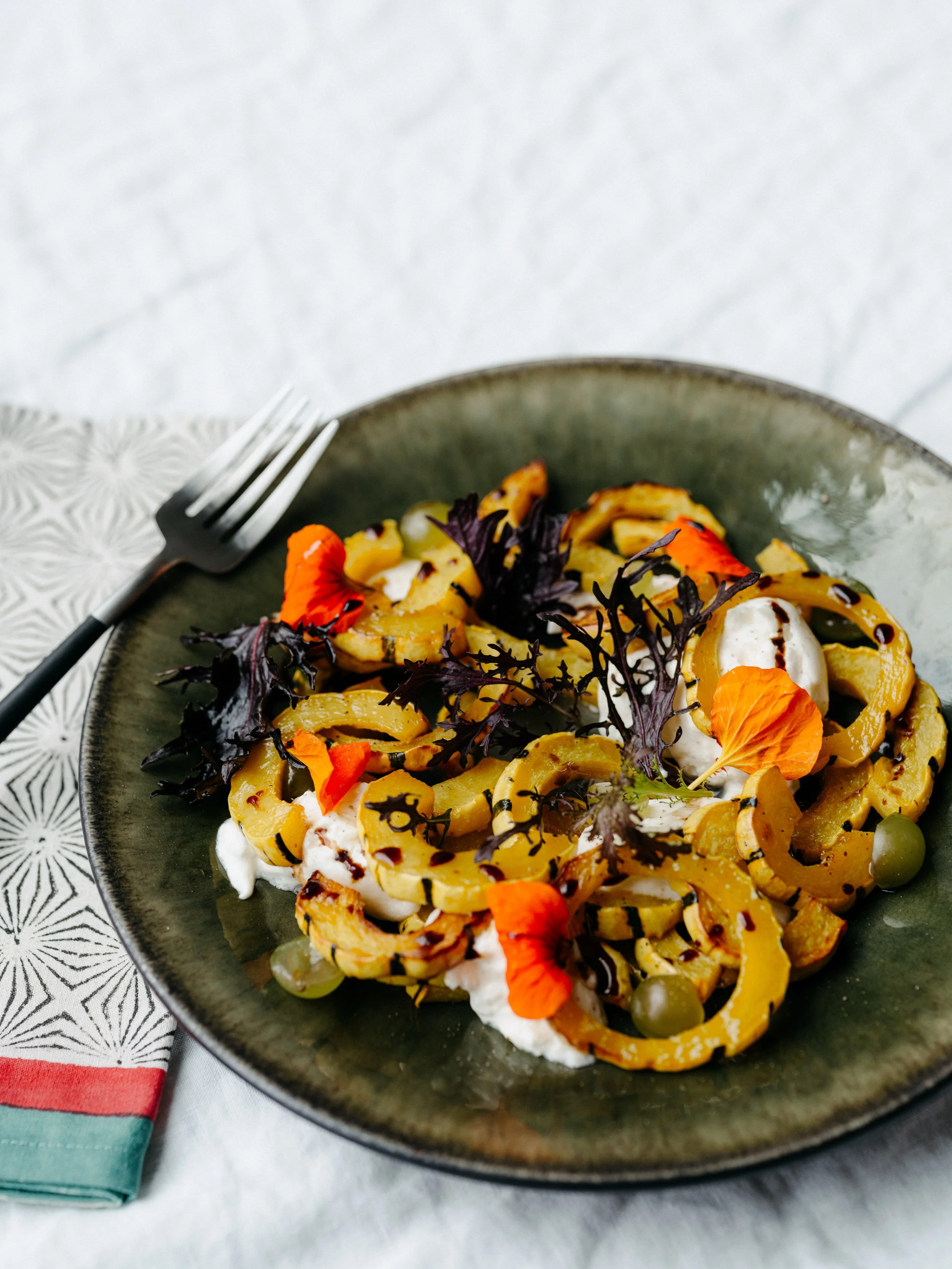 A plate of pasta garnished with edible flowers, dark leafy greens, green peas, and a drizzle of sauce, served with a fork to the side.