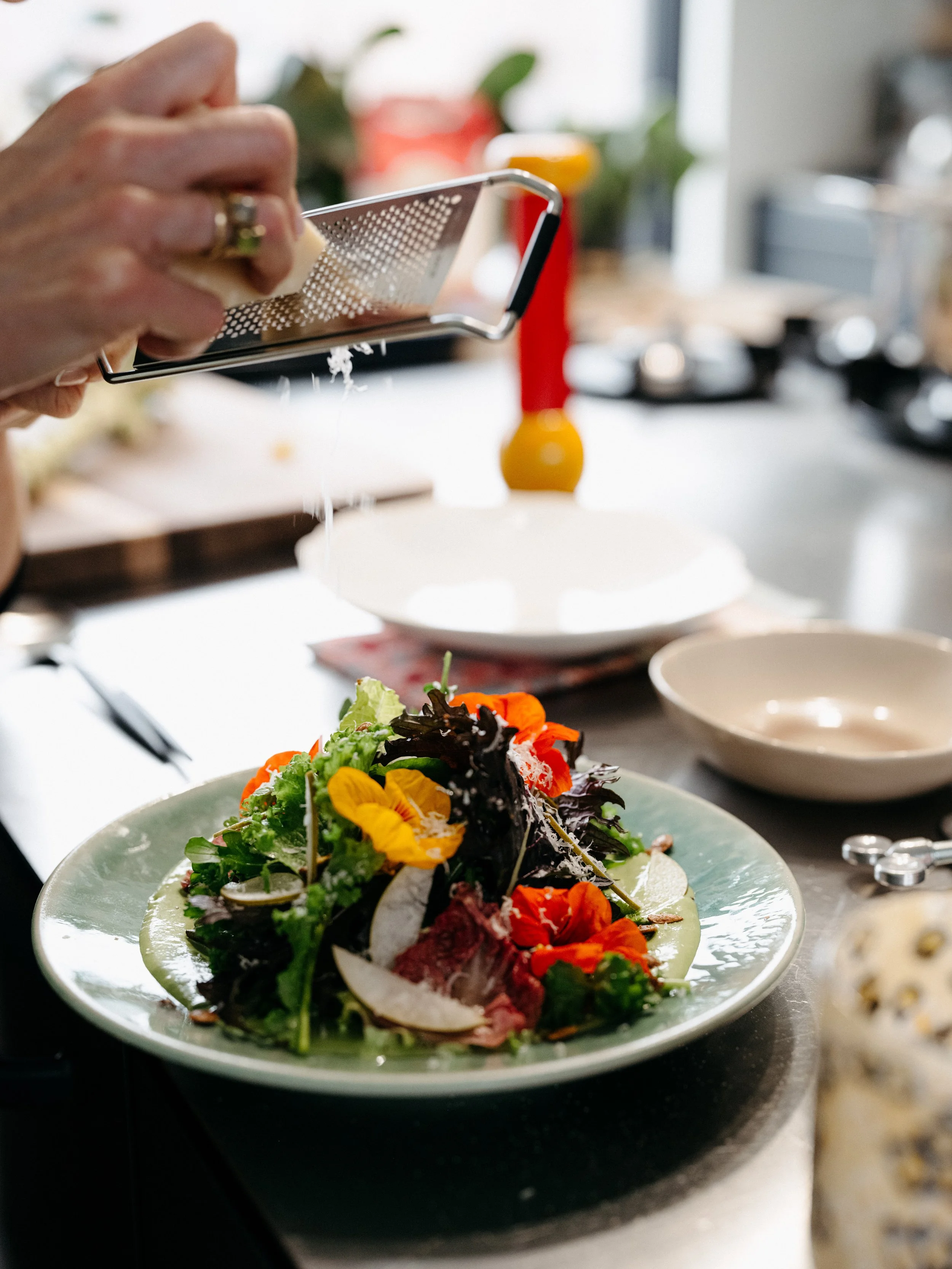 Person grating cheese over a fresh salad with mixed greens, edible flowers, and vegetables on a plate in a kitchen.