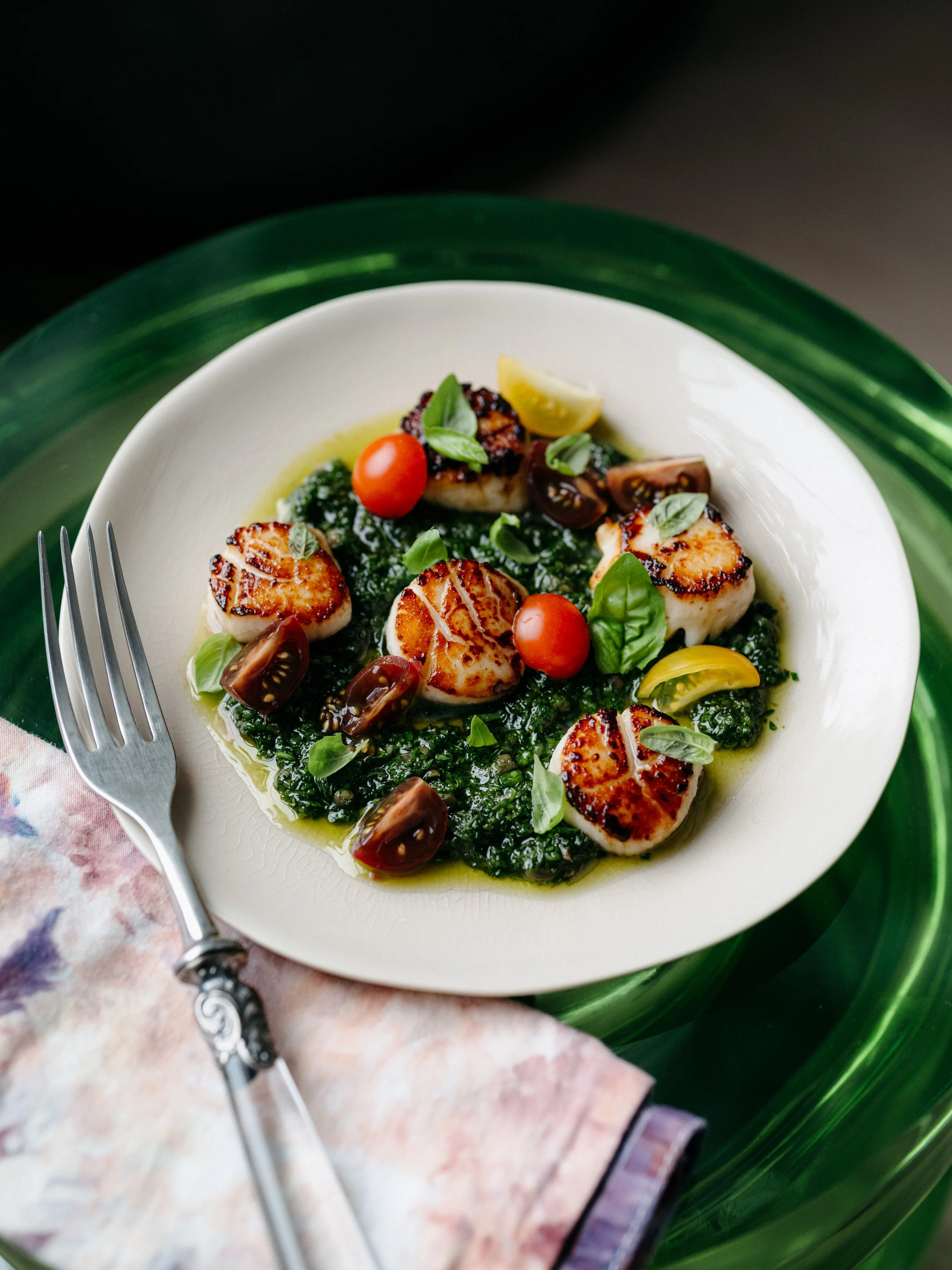 Seared scallops on a bed of sautéed greens topped with cherry tomatoes, basil, and lemon wedges, served on a white plate with a fork and a floral napkin.