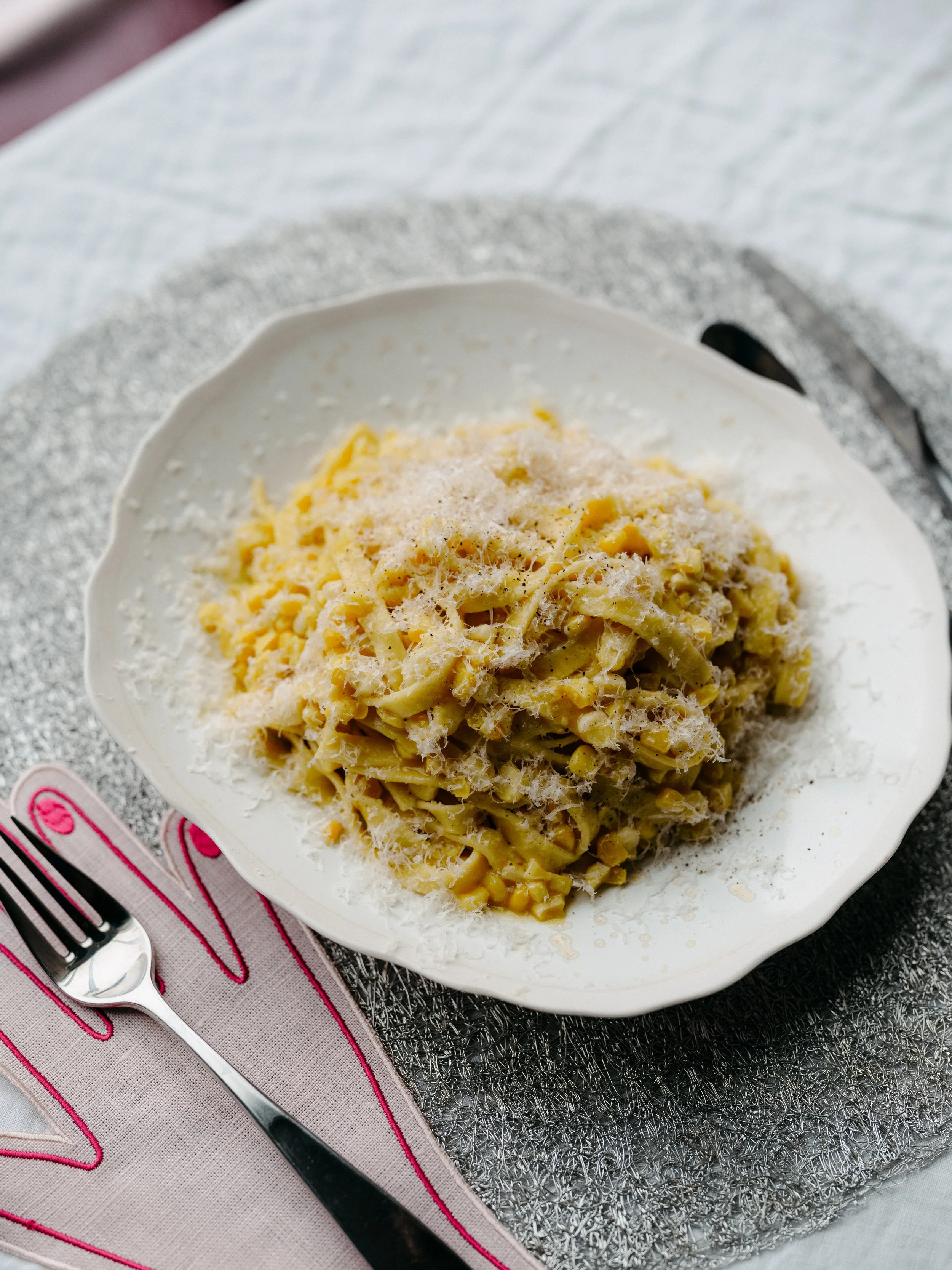 A white bowl filled with cooked fettucine pasta topped with grated cheese, placed on a decorative placemat with a fork and a knife beside it.