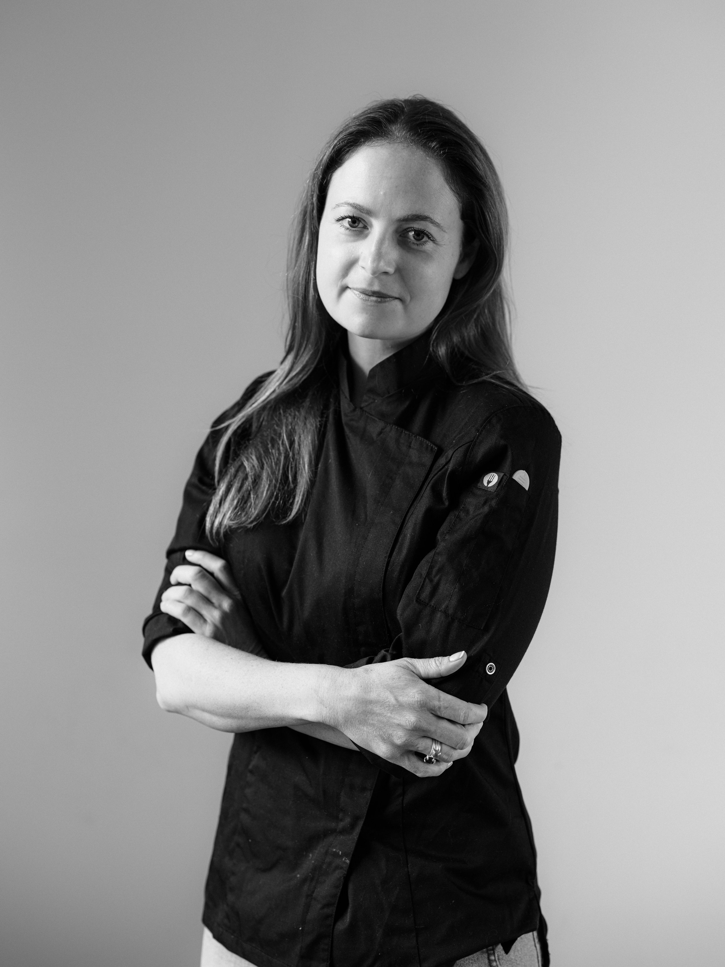 Black and white portrait of a woman with long hair, wearing a chef's coat, standing with her arms crossed, looking at the camera with a slight smile.