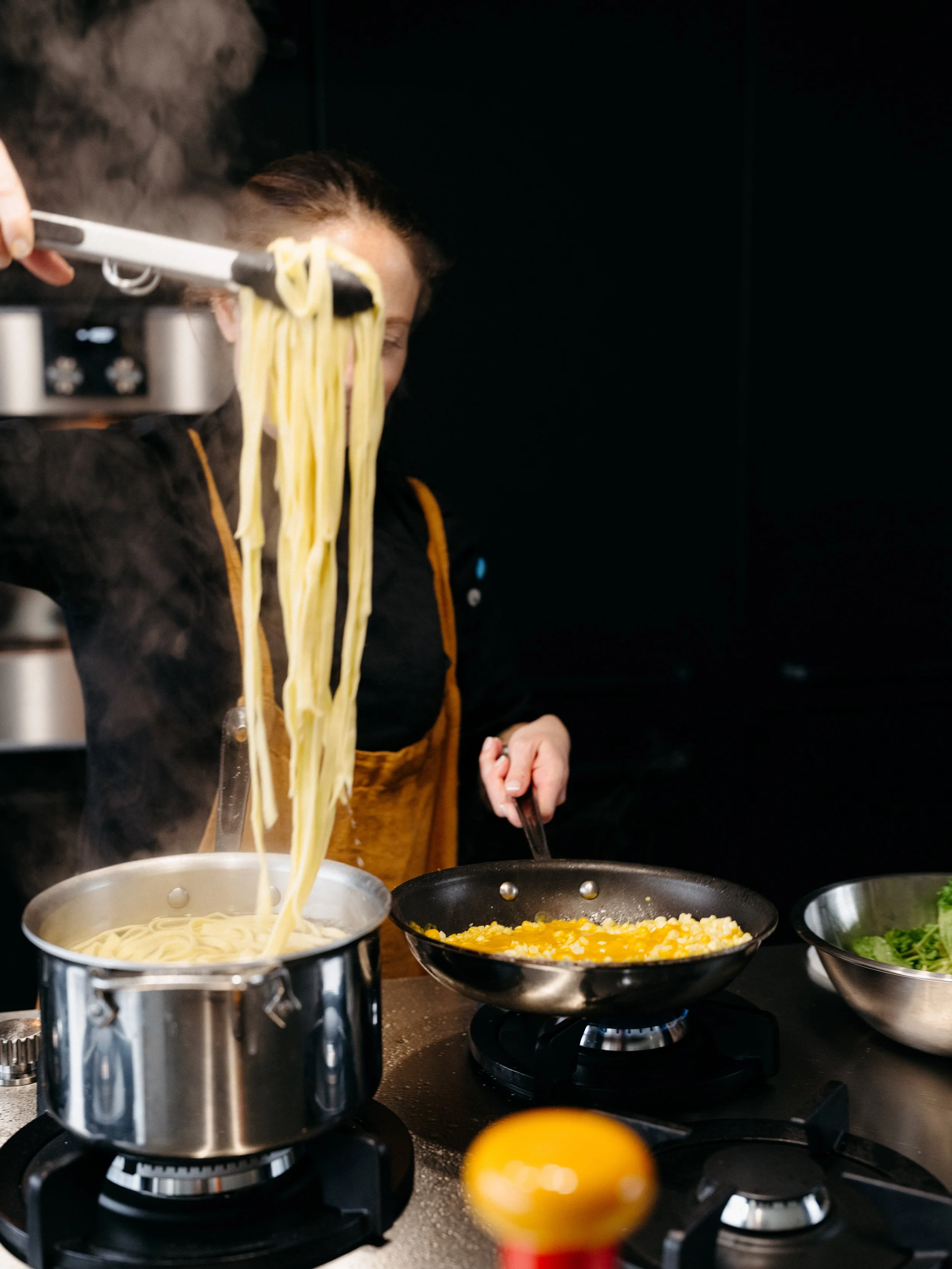 Person cooking pasta and scrambled eggs on stovetops with bowls of salad nearby.