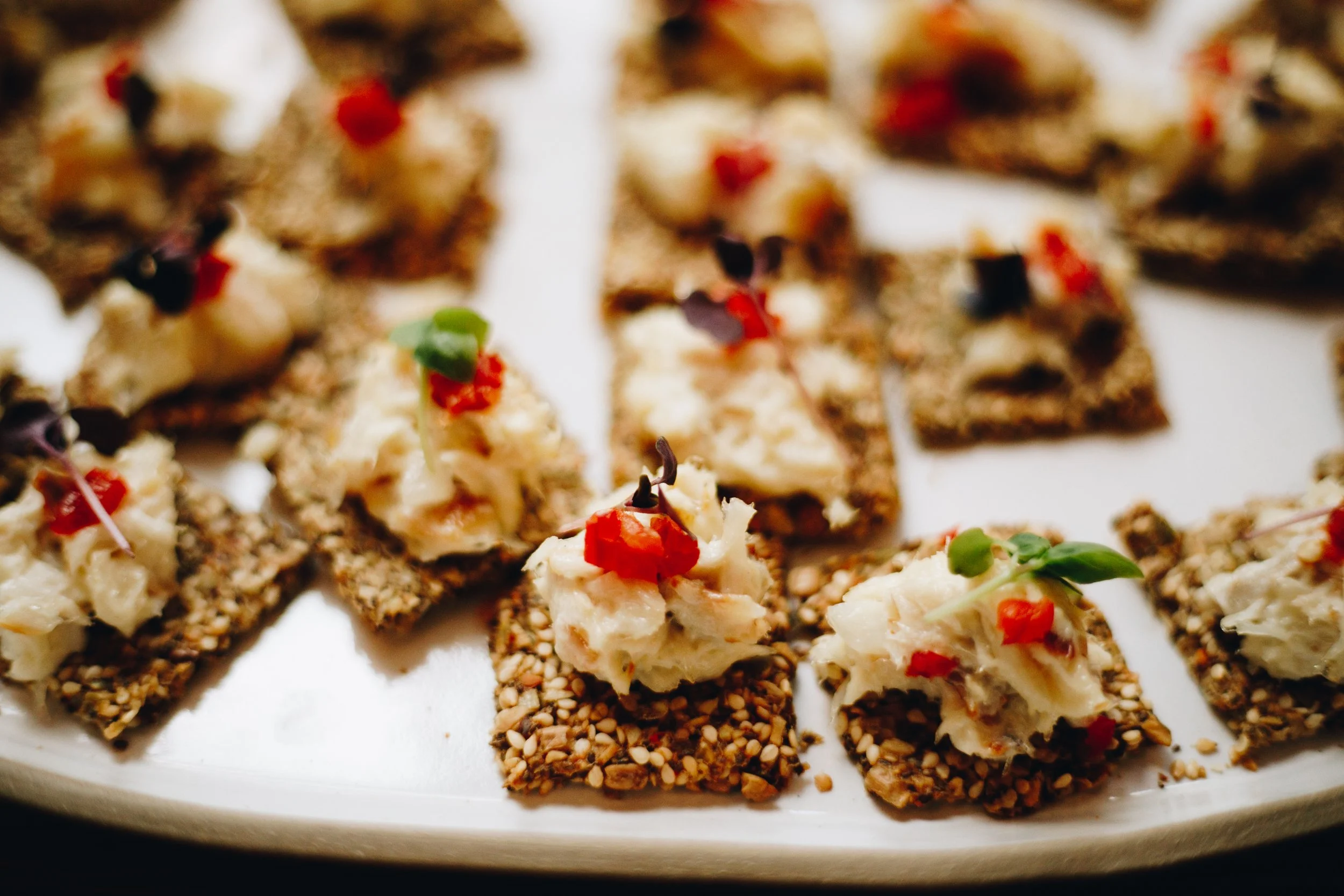 Close-up of appetizer bites with a seed-covered cracker base topped with creamy cheese, small pieces of red and green peppers, and garnished with microgreens, arranged on a white serving platter.